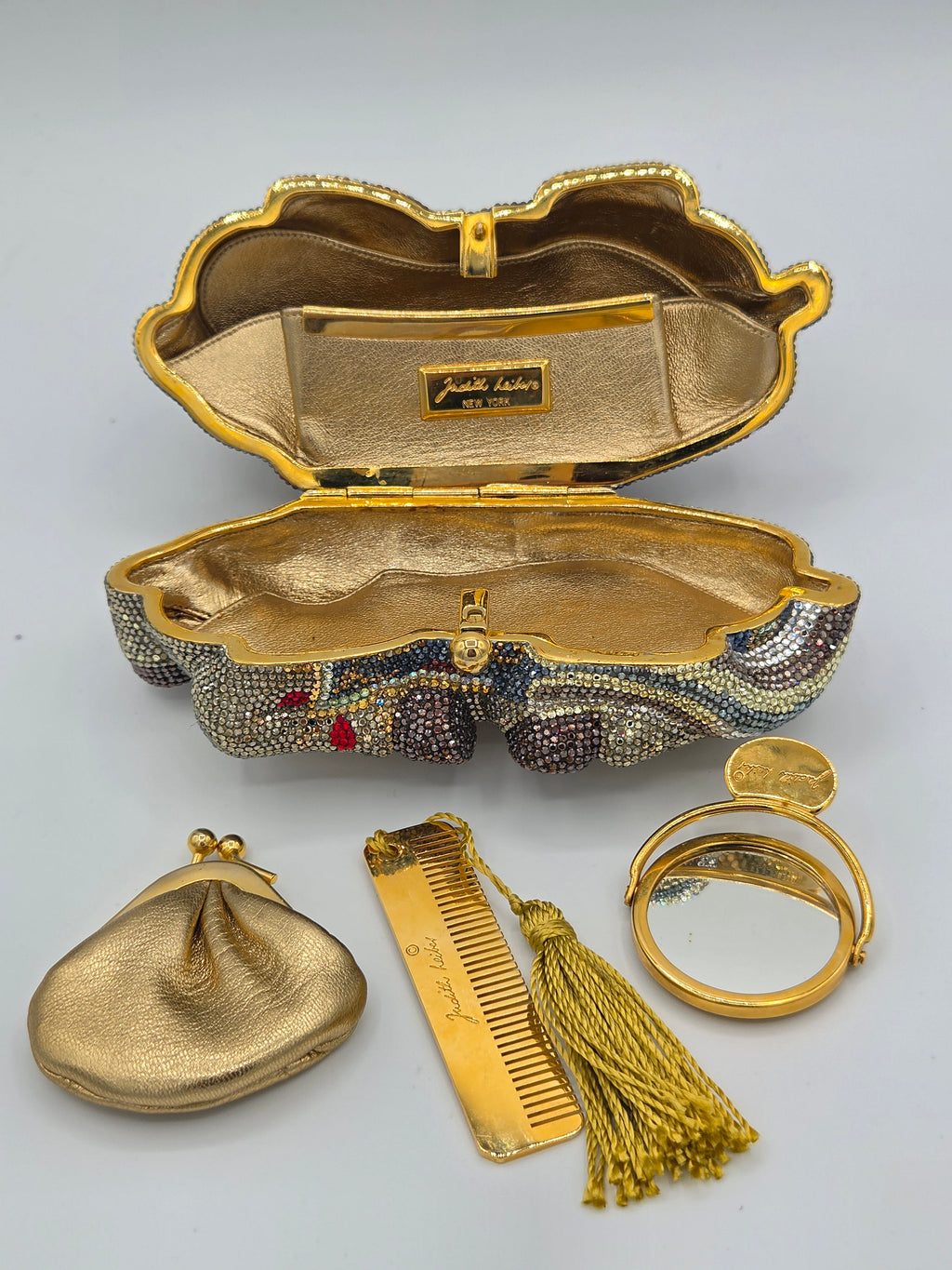Gold clutch with embellishments, a small round mirror, a gold comb, and a decorative tassel on a light gray background.