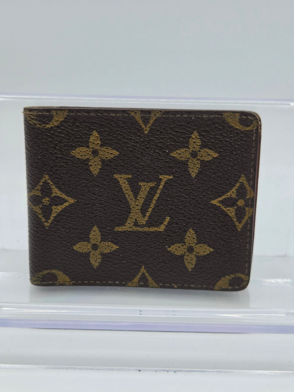 A Louis Vuitton monogram canvas passport cover or small wallet in dark brown with gold-tone printed monogram motifs, photographed inside a clear plastic sleeve against a white background.