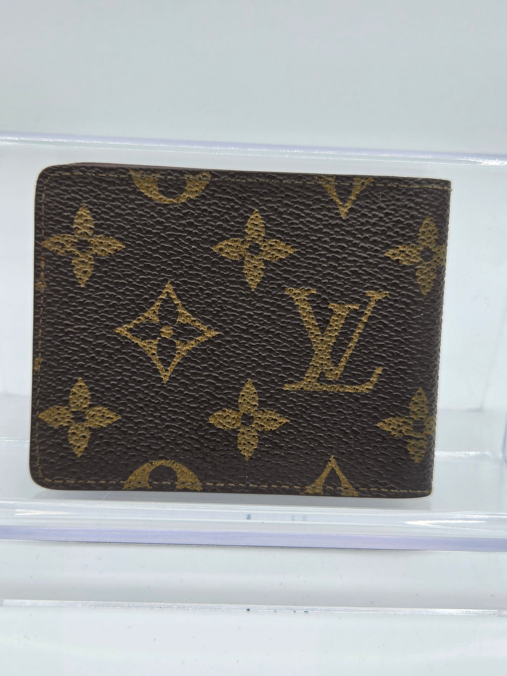 A Louis Vuitton monogram bifold wallet in brown coated canvas with gold-tone printed monogram motifs, displayed on a clear acrylic stand against a white background.