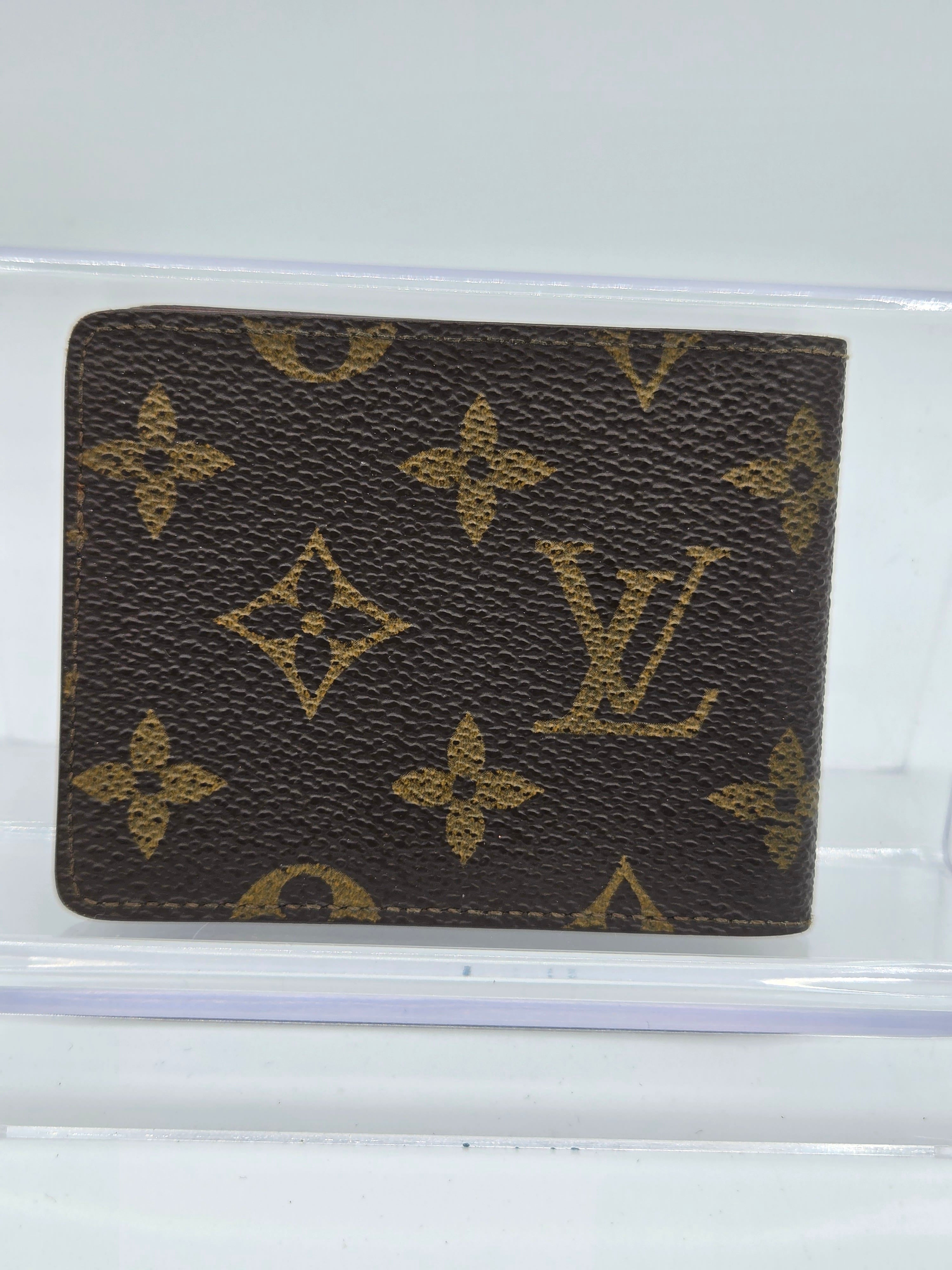 A Louis Vuitton monogram bifold wallet in brown coated canvas with gold-tone printed monogram motifs, displayed on a clear acrylic stand against a white background.