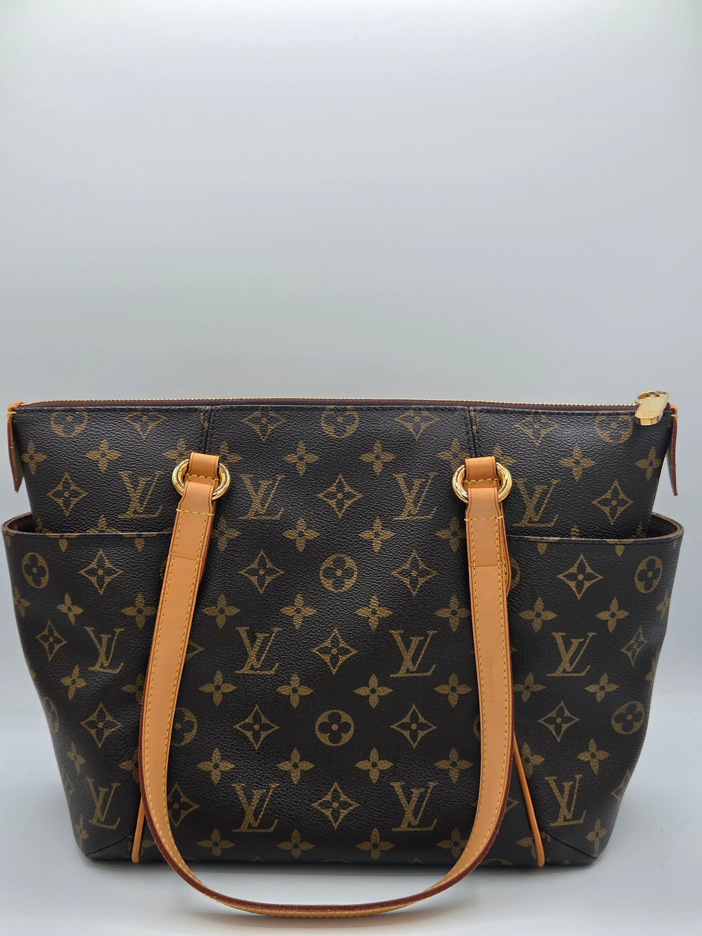A Louis Vuitton monogram canvas tote bag with tan leather handles and gold-tone hardware, shown from a side angle against a light gray background.