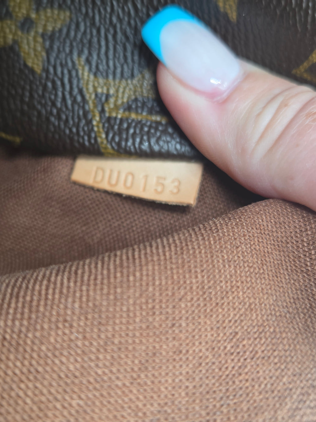Close-up of a Louis Vuitton monogram bag with a person's finger touching the brown canvas lining, showing a small tan leather tag with the number DUO153.