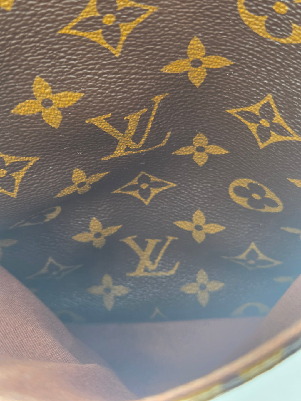 Close-up of a brown monogram fabric with gold Louis Vuitton-style floral and LV motifs, showing part of a curved brown object edge.