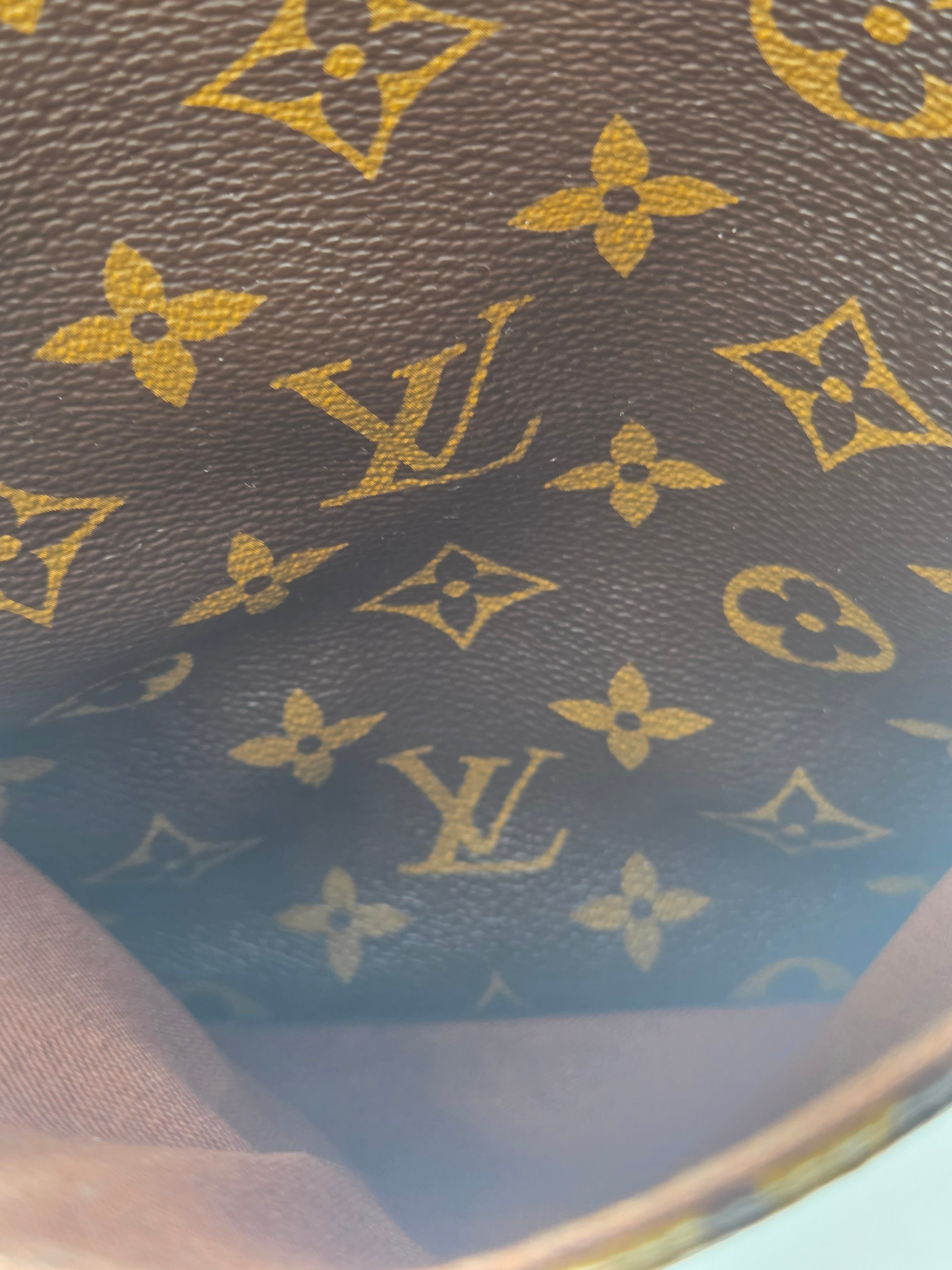 Close-up of a brown monogram fabric with gold Louis Vuitton-style floral and LV motifs, showing part of a curved brown object edge.