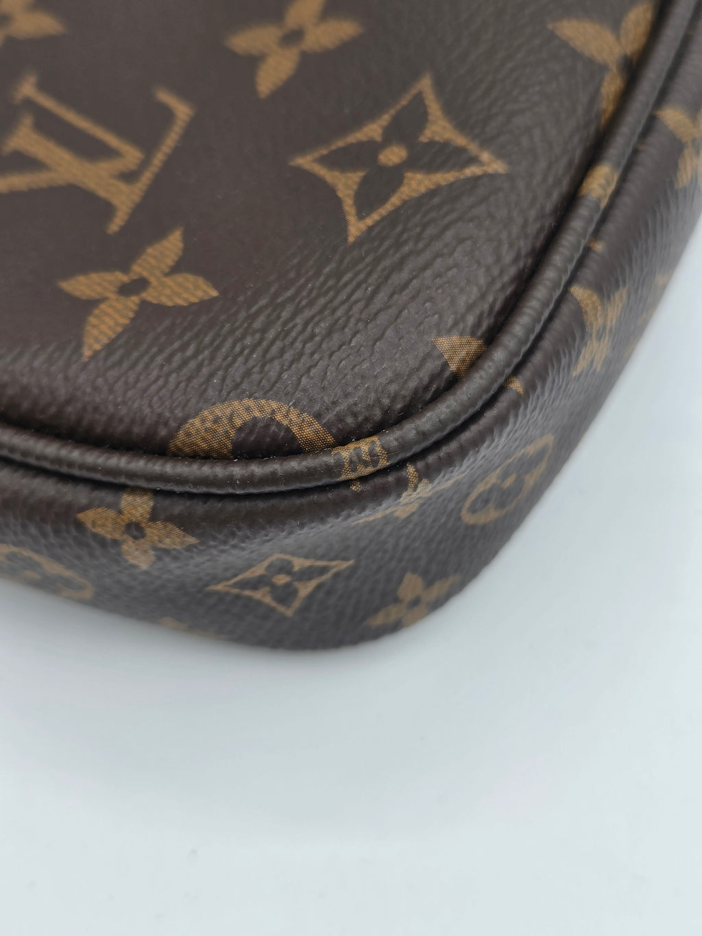 Close-up of a brown coated canvas item featuring the classic tan Louis Vuitton monogram pattern, showing the textured leather grain and a curved seam against a plain light background.