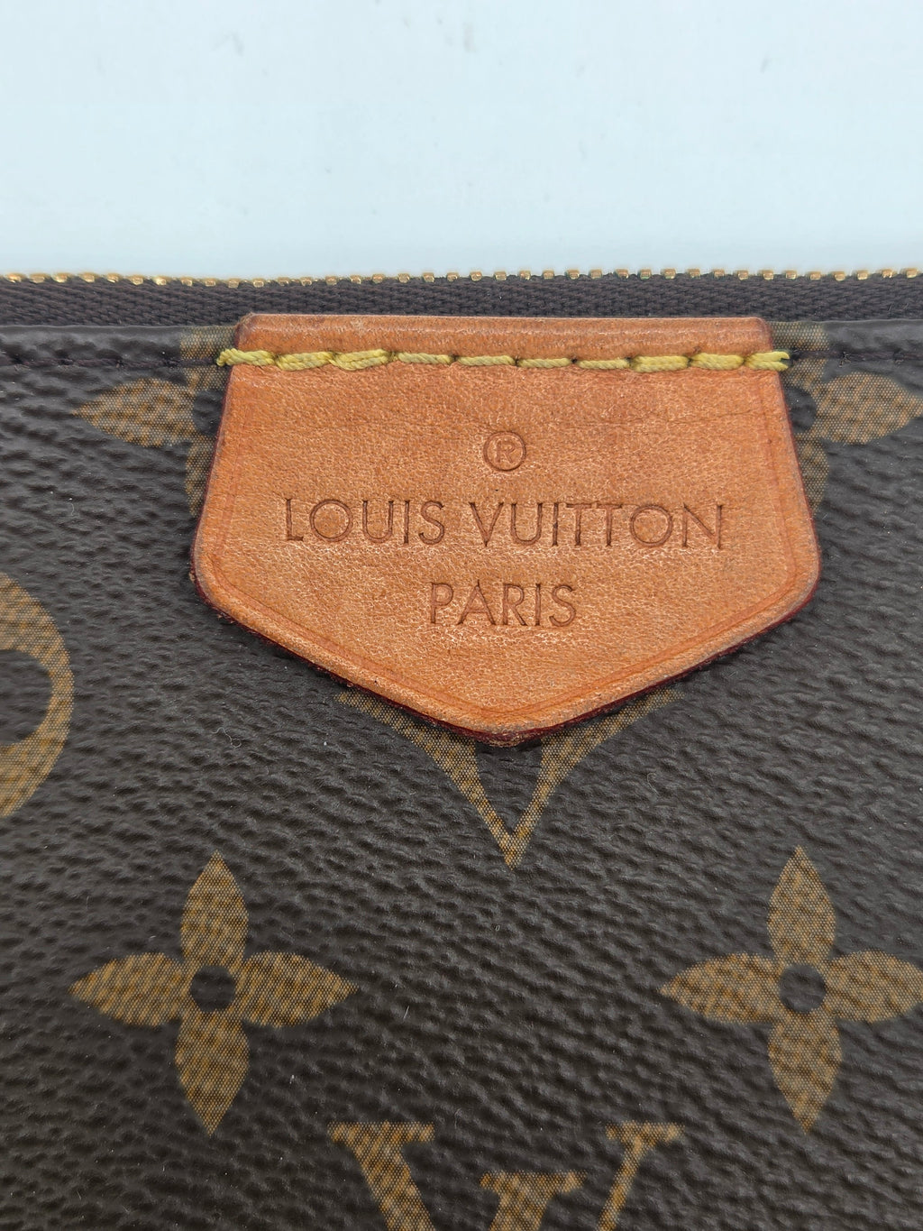 Close-up of a Louis Vuitton monogram canvas item with a tan leather tag embossed with 'LOUIS VUITTON PARIS' and gold-tone stitching.