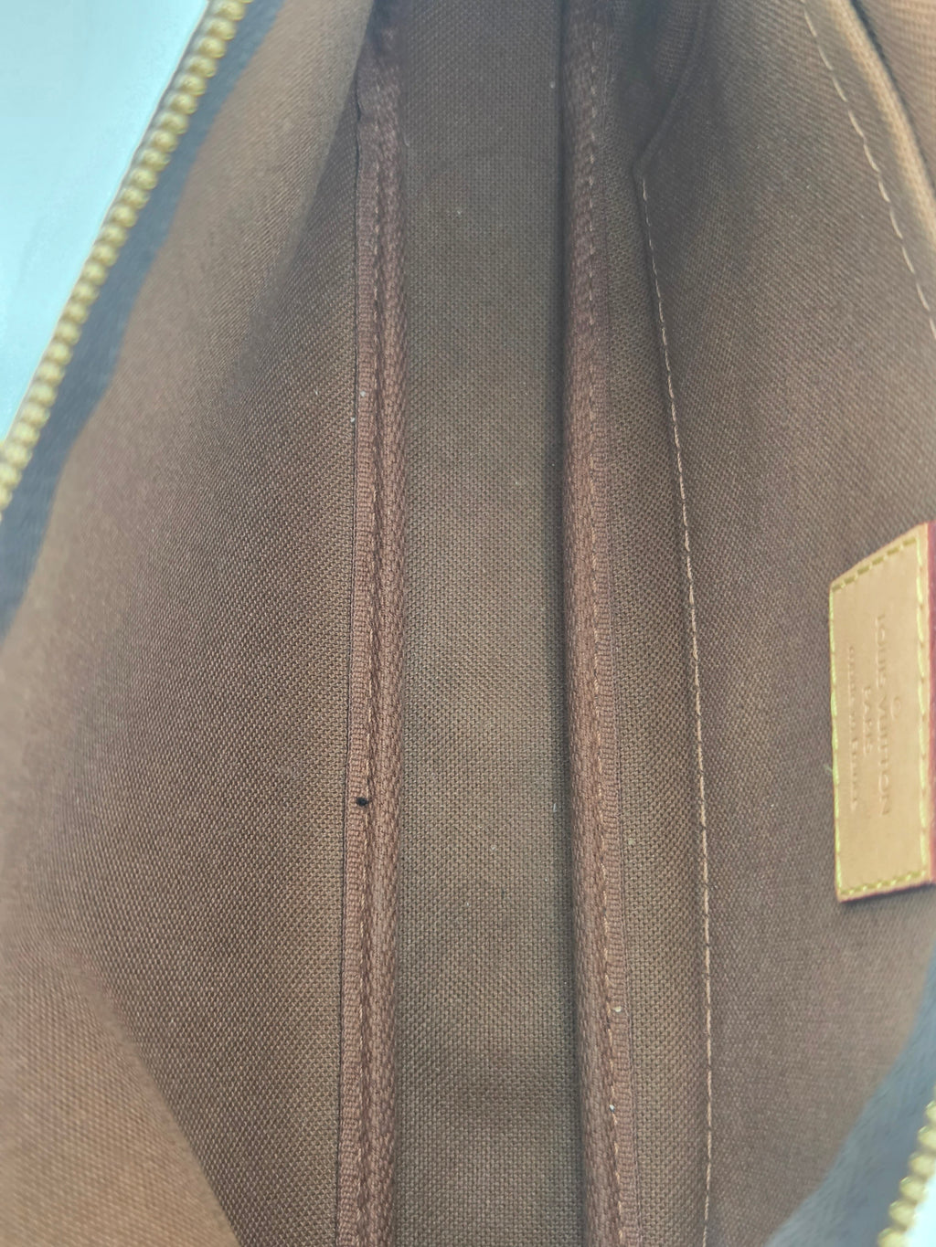 Close-up of the interior lining of a Louis Vuitton bag, showing a brown canvas body, double-stitched compartments, and a gold-tone zipper with a small beige brand patch at the top.