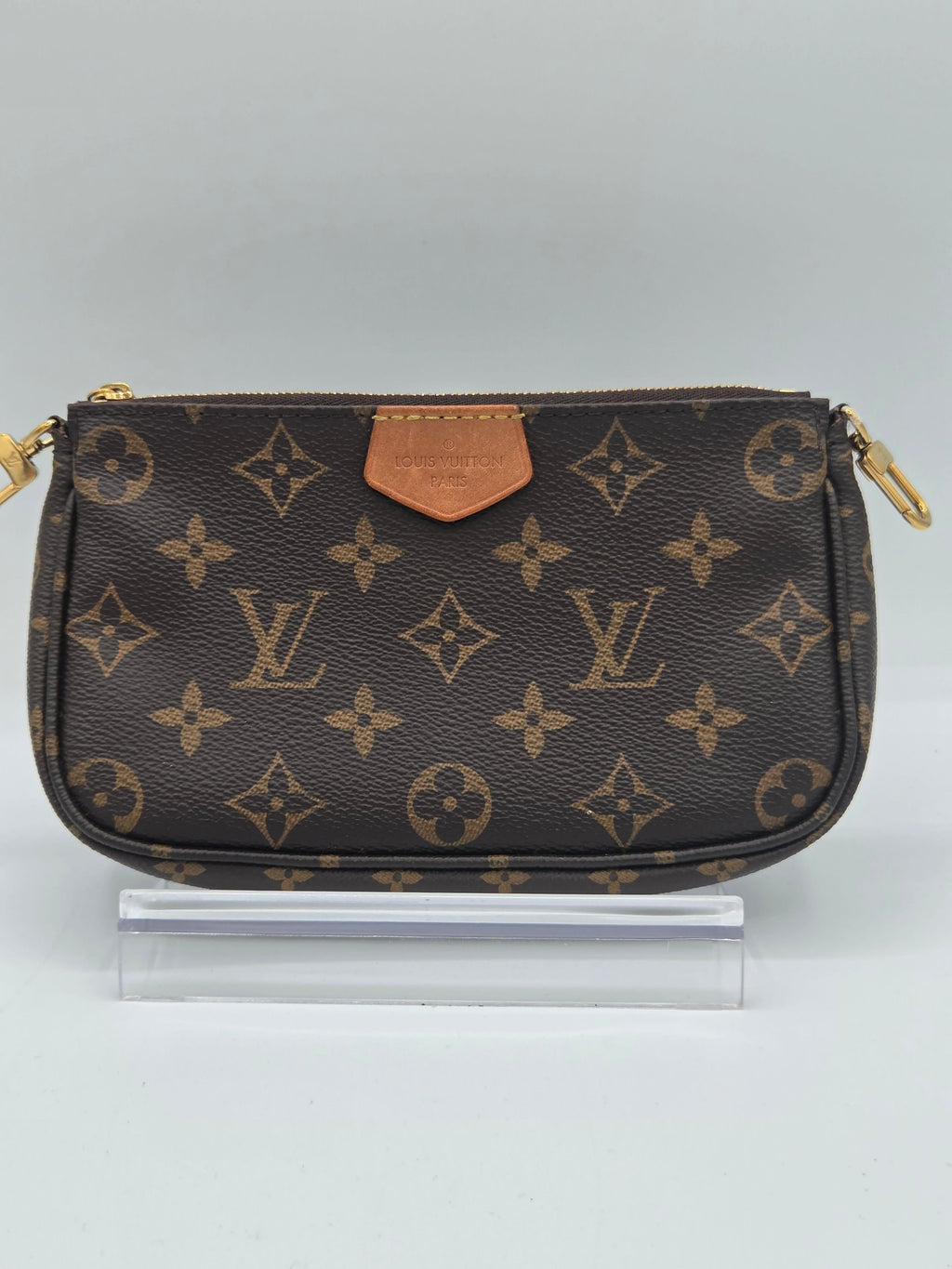 A Louis Vuitton monogram canvas pouch in dark brown with gold-tone hardware and a tan leather accent, shown next to a clear plastic stand on a light gray background.