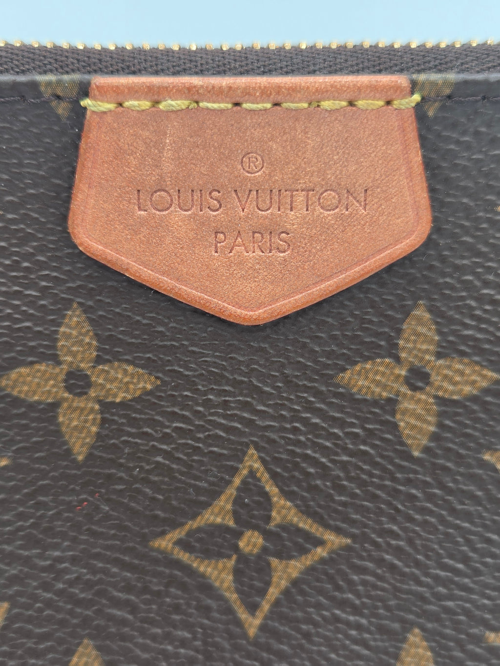 Close-up of a Louis Vuitton monogram coated canvas bag, showing the dark brown canvas with tan/gold monogram pattern and a tan leather brand tab embossed with 'LOUIS VUITTON PARIS'.