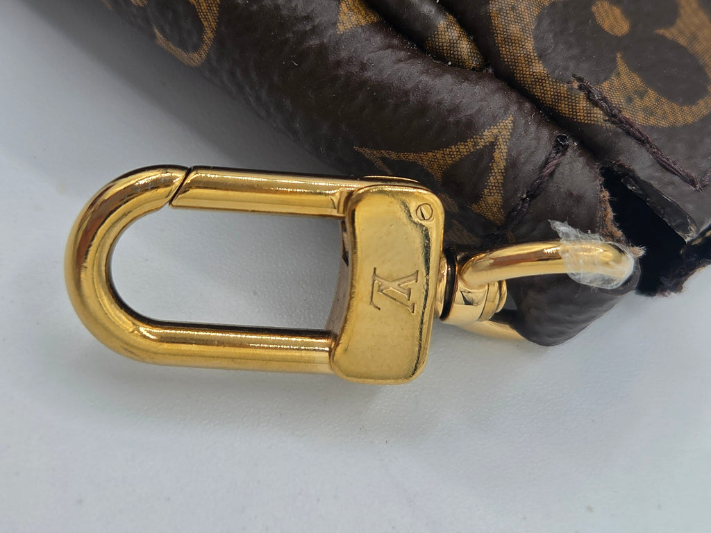 A close-up of a Louis Vuitton monogram bag's gold-tone metal swivel clasp and attachment ring, attached to a dark brown and gold patterned fabric.