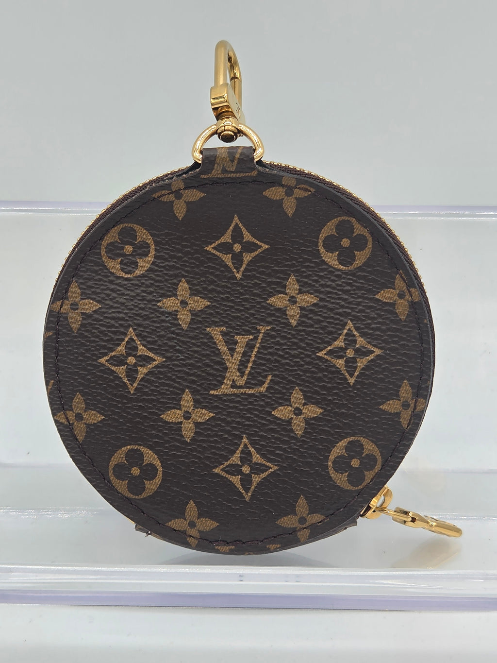 A Louis Vuitton monogram canvas round coin pouch with gold-tone hardware, shown hanging against a light gray background.