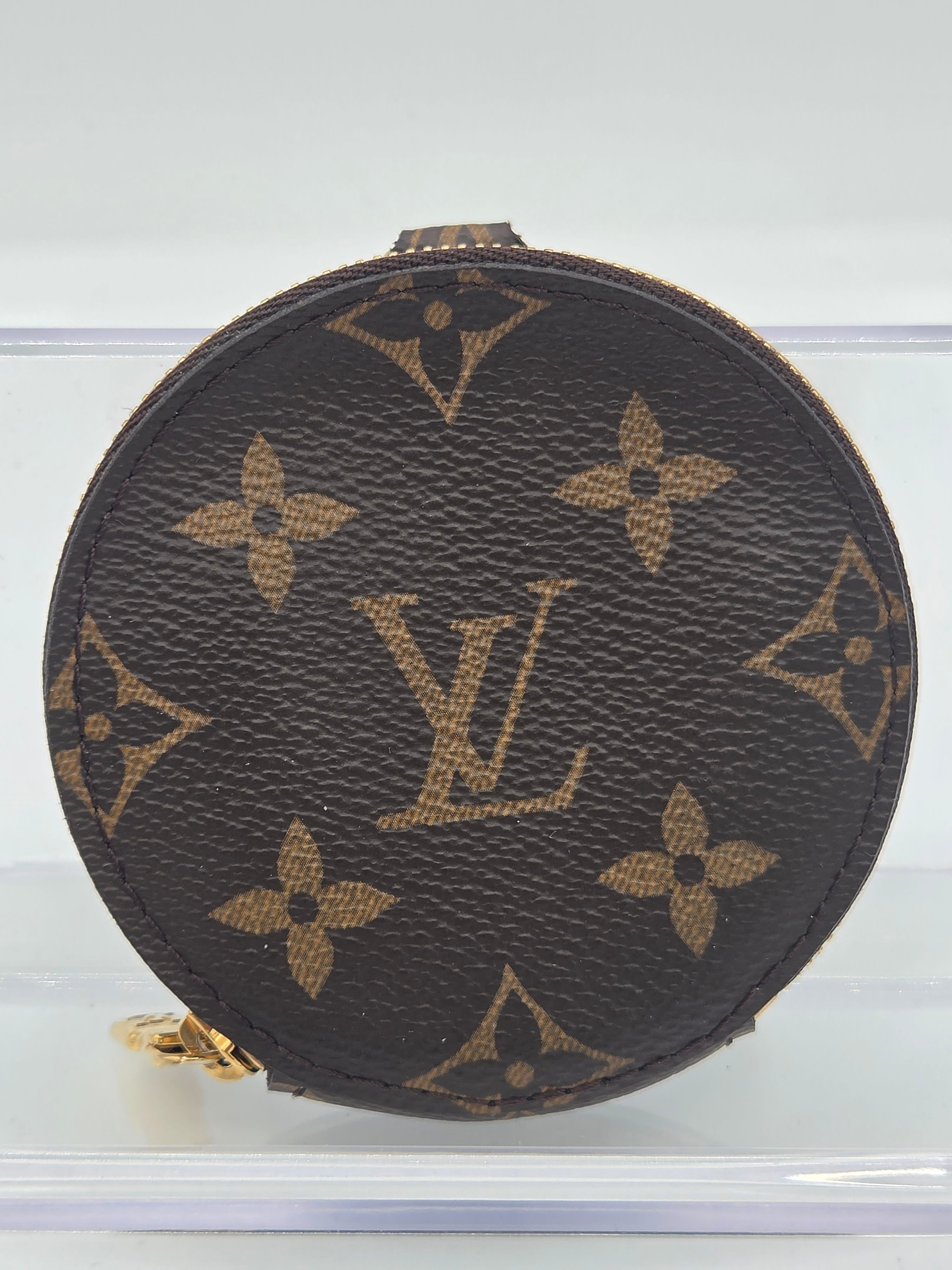 A round Louis Vuitton coin pouch in brown monogram canvas with gold-tone hardware, shown against a light background.