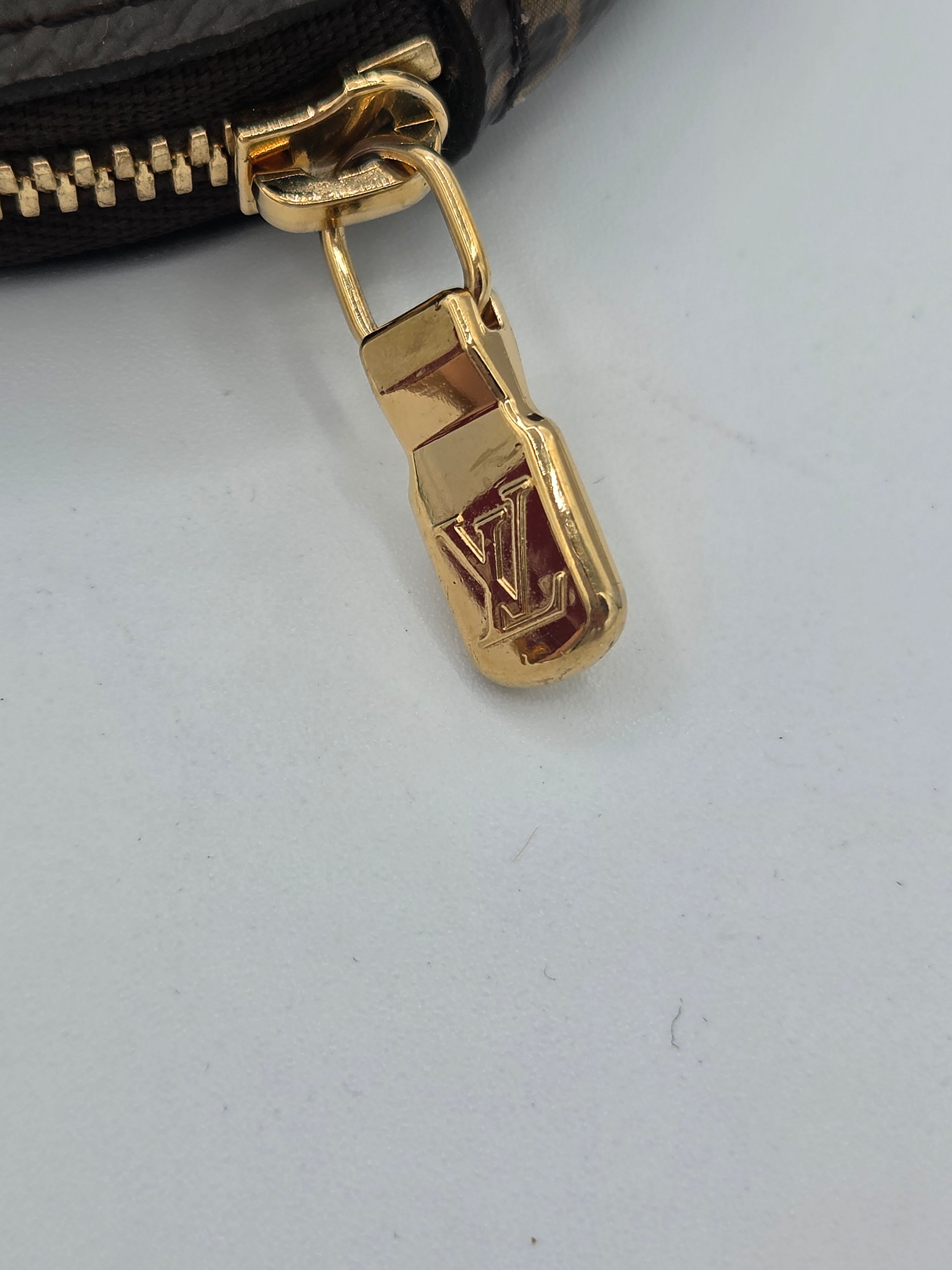 Close-up of a gold-tone metal zipper pull with an embossed LV logo, attached to a black zipper on a light gray background.