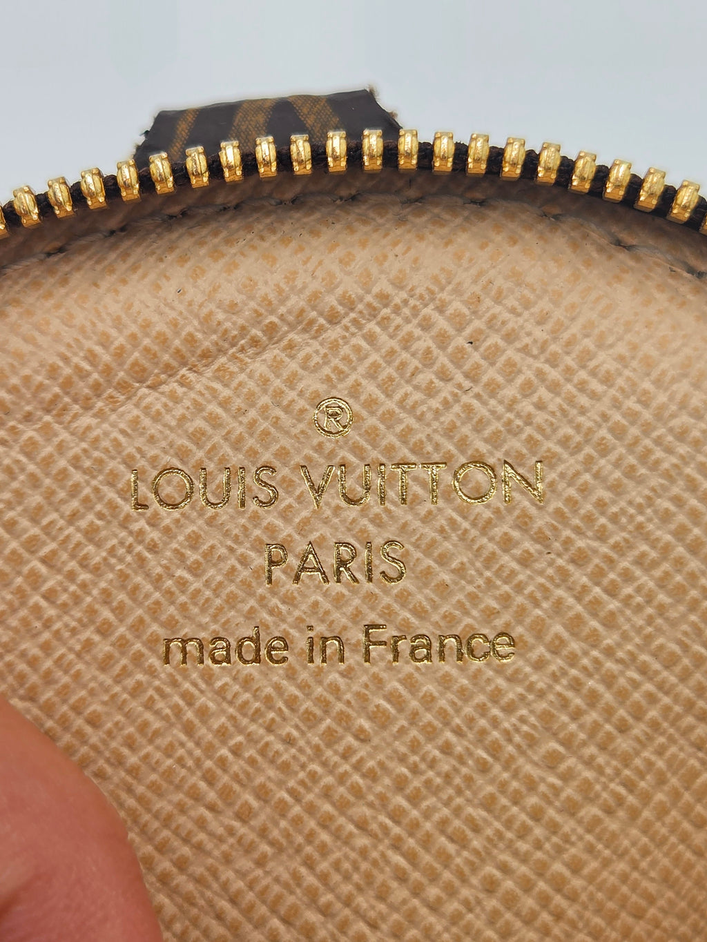 Close-up of a Louis Vuitton pouch in beige Saffiano canvas with gold-toned zipper trim and embossed 'LOUIS VUITTON PARIS made in France' text, held by a hand.
