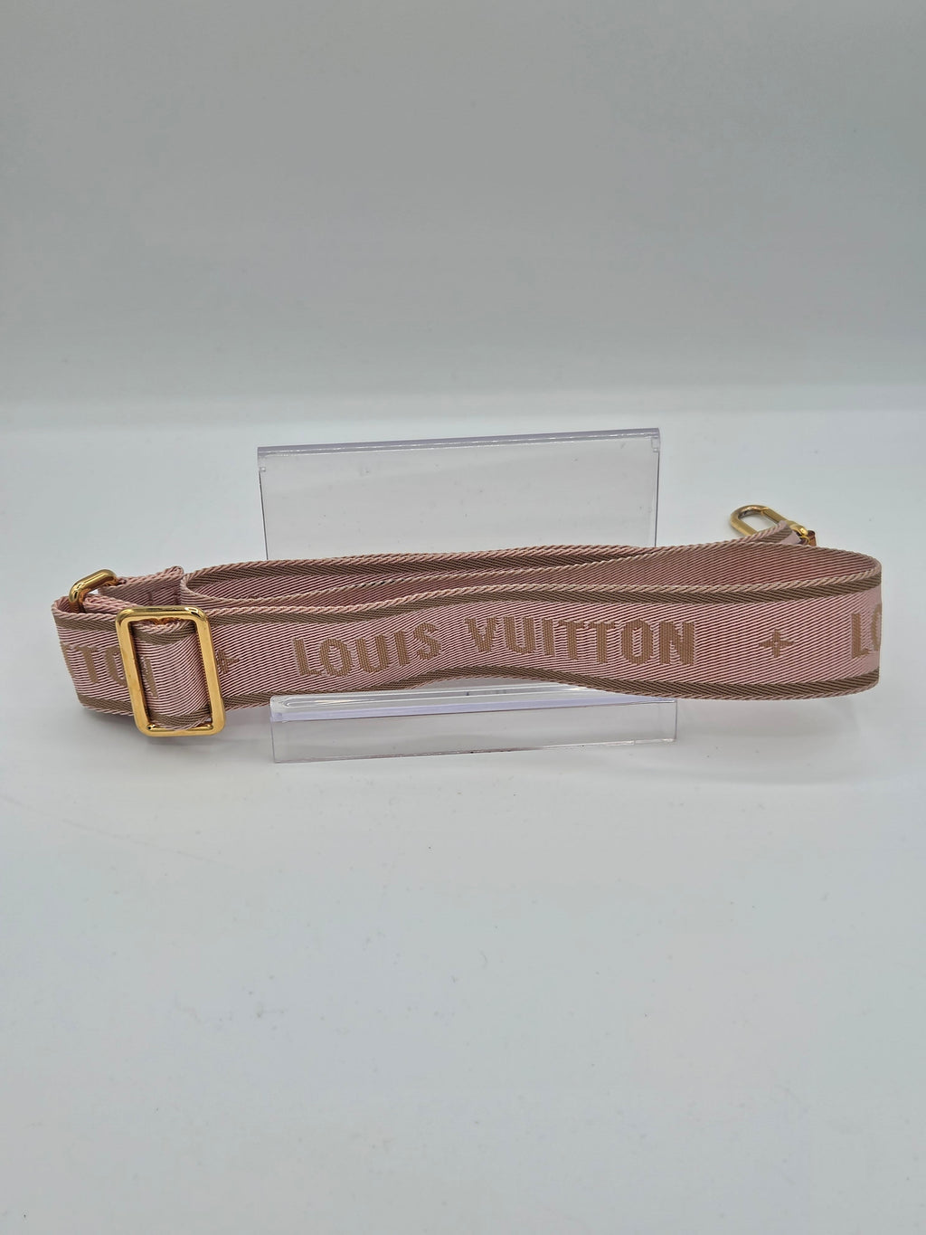 A Louis Vuitton shoulder strap in a dusty rose pink color with gold-tone hardware, displayed on a clear acrylic stand against a plain white background.