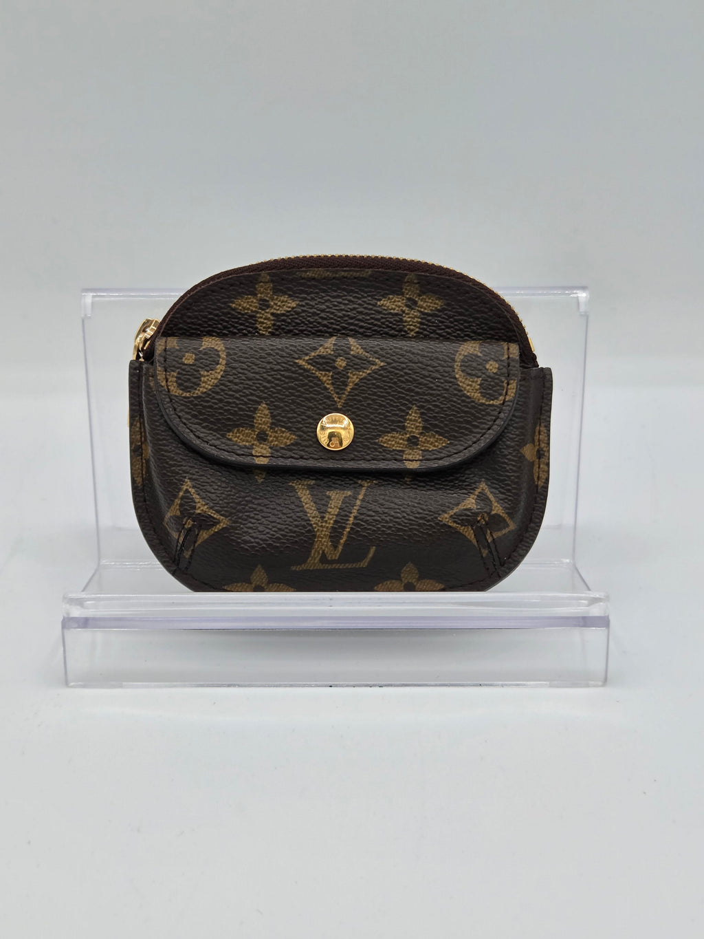 A Louis Vuitton monogram canvas coin pouch with a gold-tone snap button, displayed inside a clear acrylic block on a light gray background.