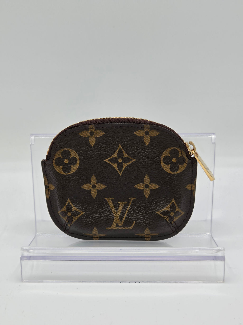 A Louis Vuitton monogram canvas coin pouch with a gold zipper, mounted on a clear acrylic display stand against a light gray background.