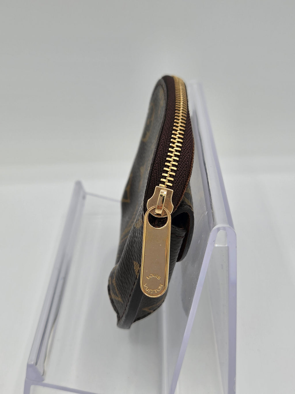 A Louis Vuitton monogram canvas zip pouch in dark brown, shown resting on a clear acrylic display stand against a white background.