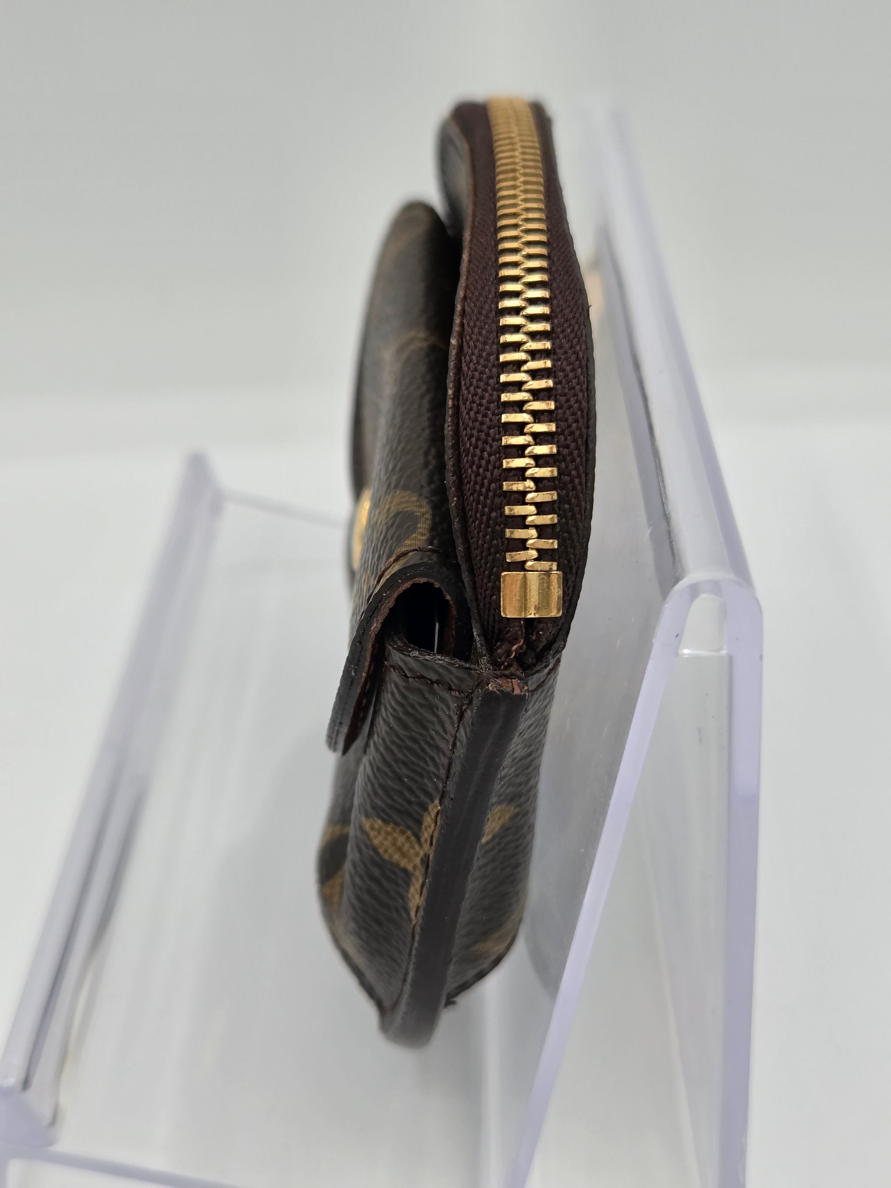 A close-up of a dark brown monogram wallet or pouch with a gold zipper, shown resting on a clear acrylic display stand against a white background.