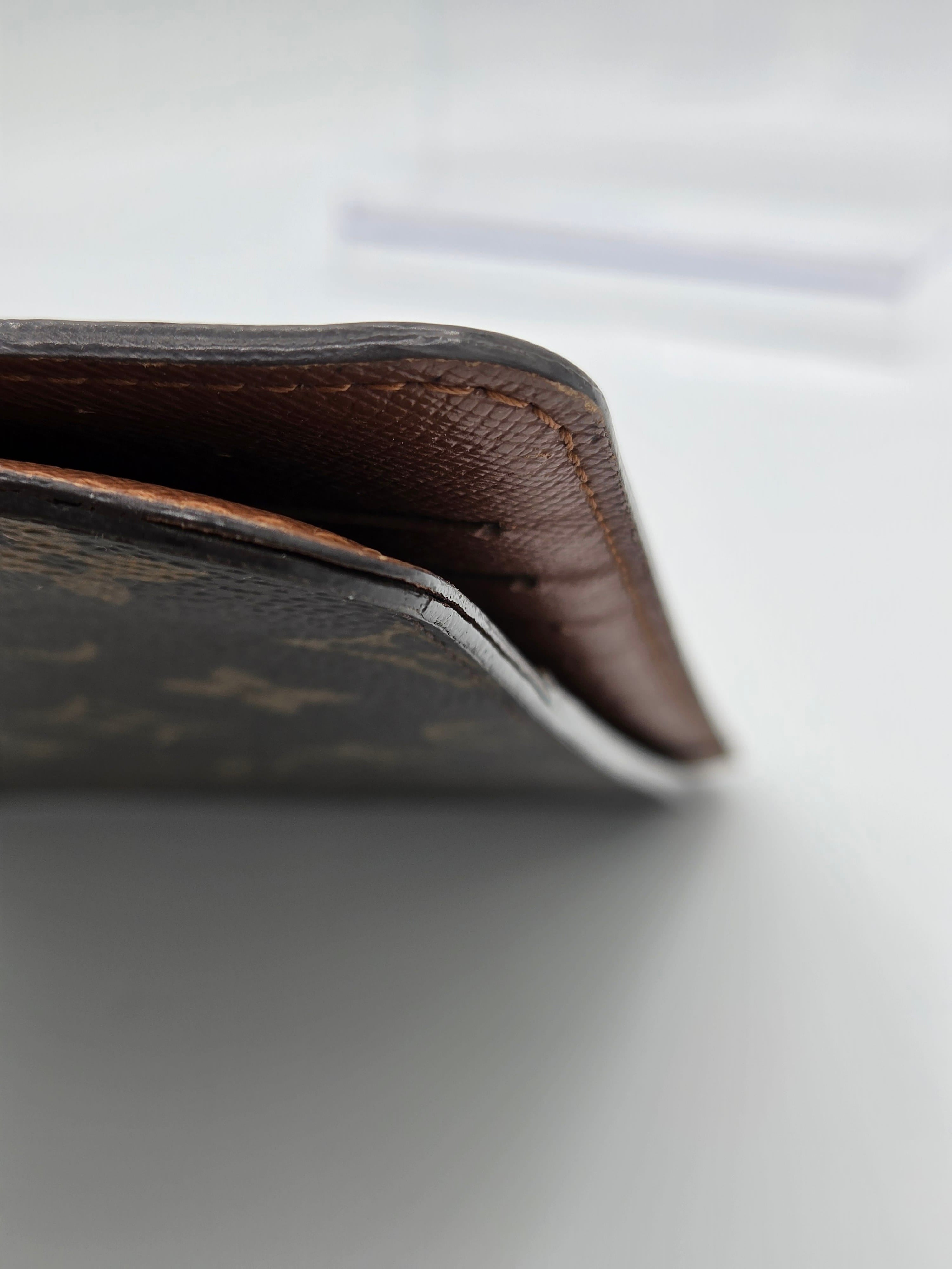 Close-up of a brown leather wallet corner with visible stitching and a dark monogram patterned exterior against a light gray background.