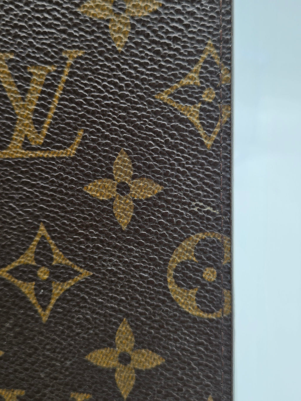 Close-up of Louis Vuitton monogram canvas with brown textured background and tan/golden LV and floral motifs, shown against a pale blue background.