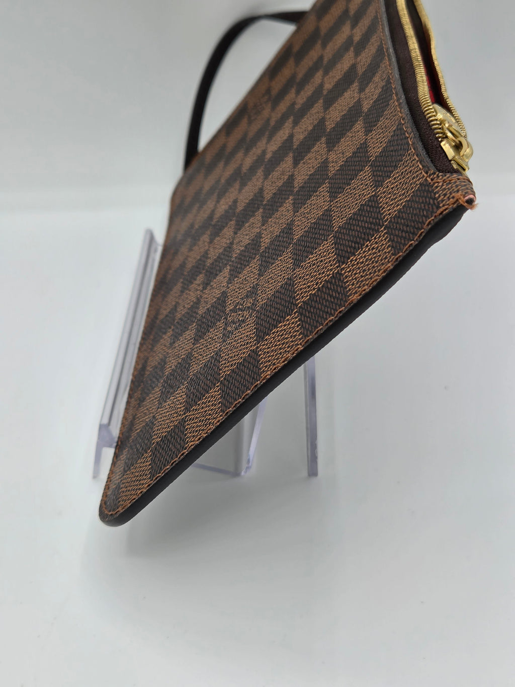 A Louis Vuitton clutch bag in the Damier Ebene checkered canvas, featuring a brown and beige pattern, gold zipper, and black leather trim, displayed on a clear acrylic stand against a white background.