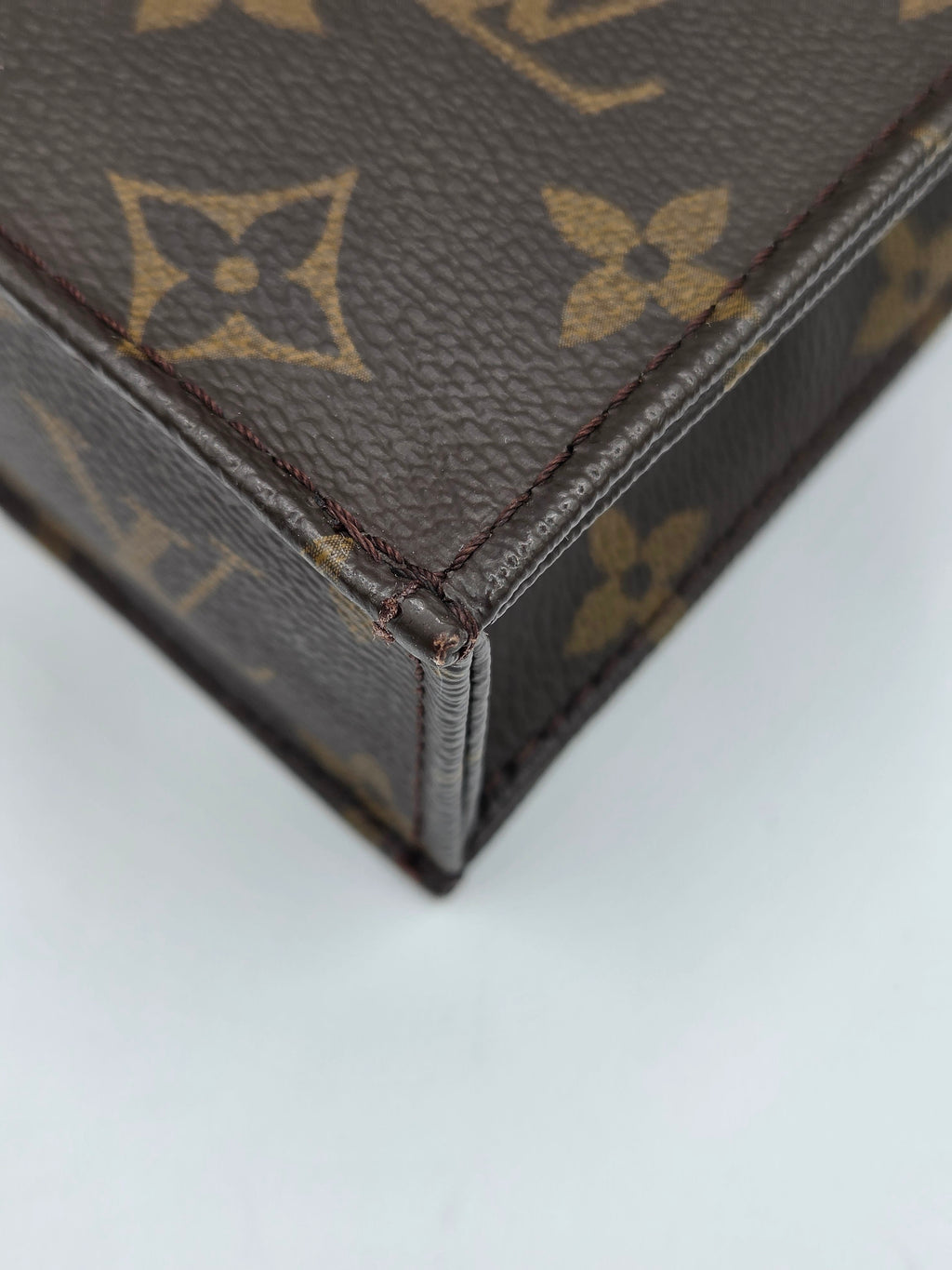 Close-up of a brown monogram canvas bag corner with tan Louis Vuitton-style floral and square motifs, showing dark brown leather trim and stitching against a light gray background.