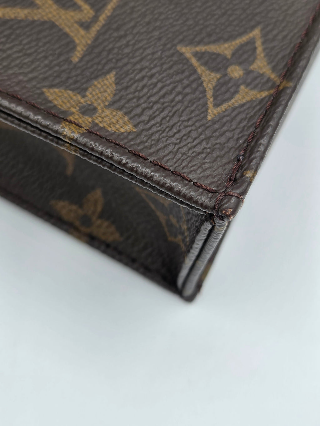 Close-up of a Louis Vuitton monogram canvas item, showing the classic brown and tan pattern and leather edge against a light gray background.
