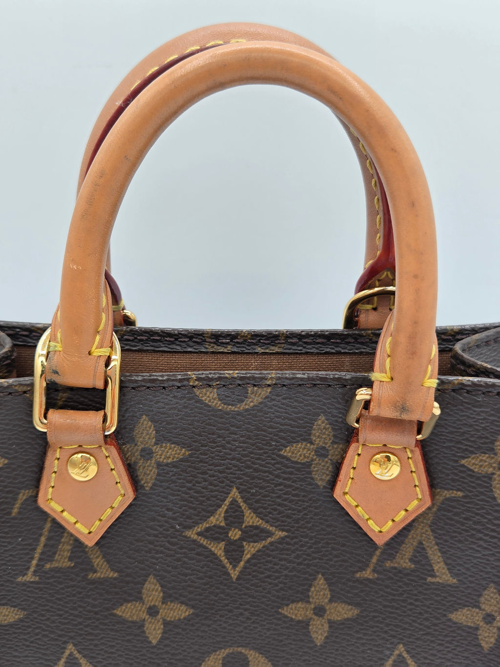 Close-up of a Louis Vuitton monogram handbag showing a tan leather handle attached with gold-tone hardware to dark brown coated canvas.