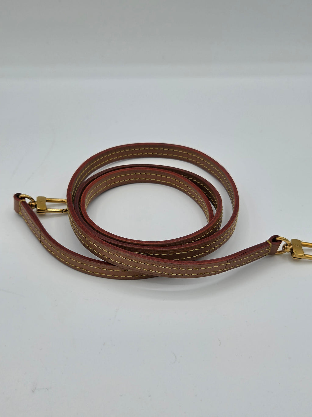 A brown leather shoulder strap with light tan contrast stitching and gold-tone metal hardware, coiled against a plain white background.