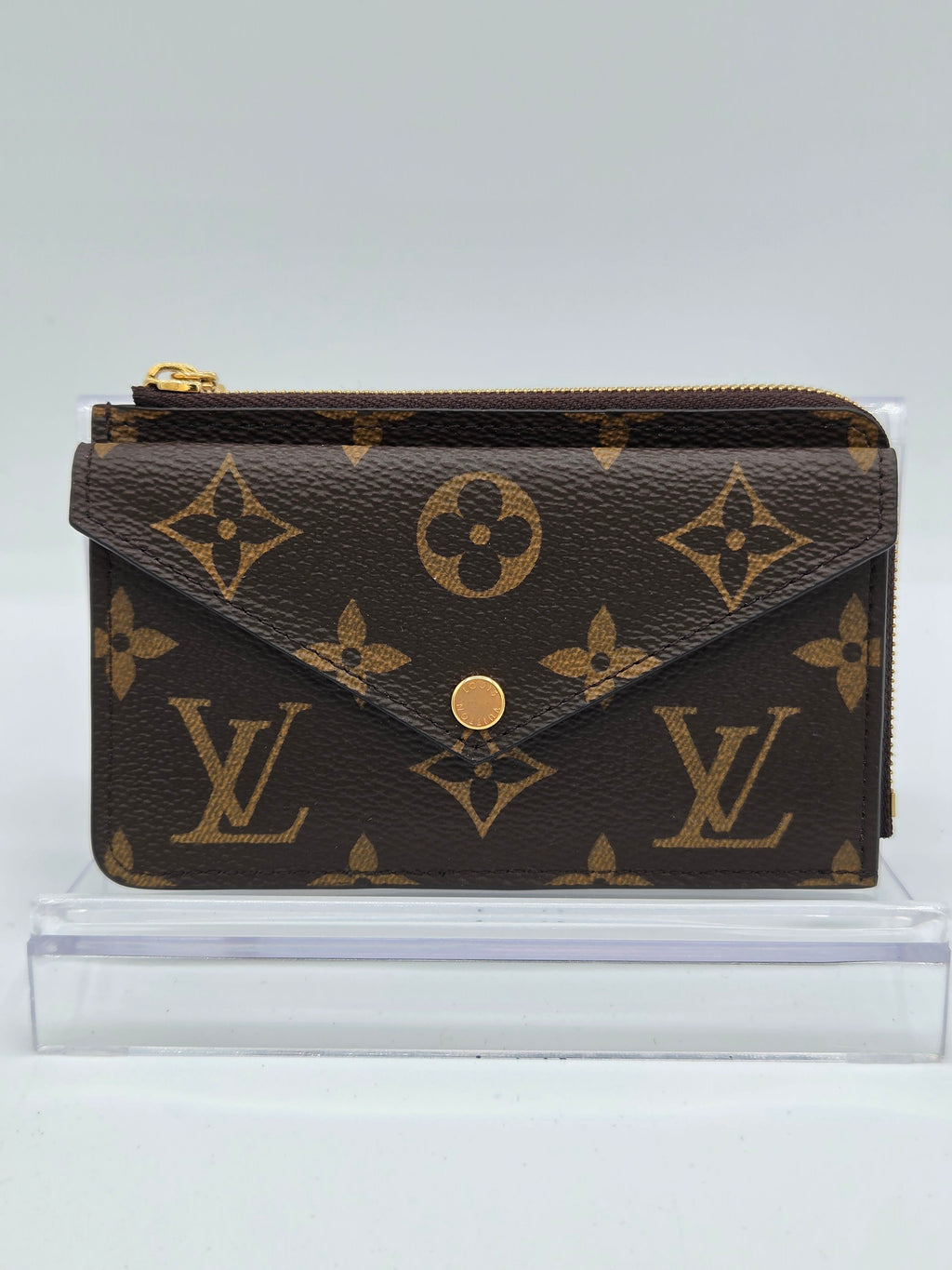 A Louis Vuitton monogram wallet in dark brown coated canvas with gold-tone hardware, shown standing upright next to a clear acrylic block against a light gray background.