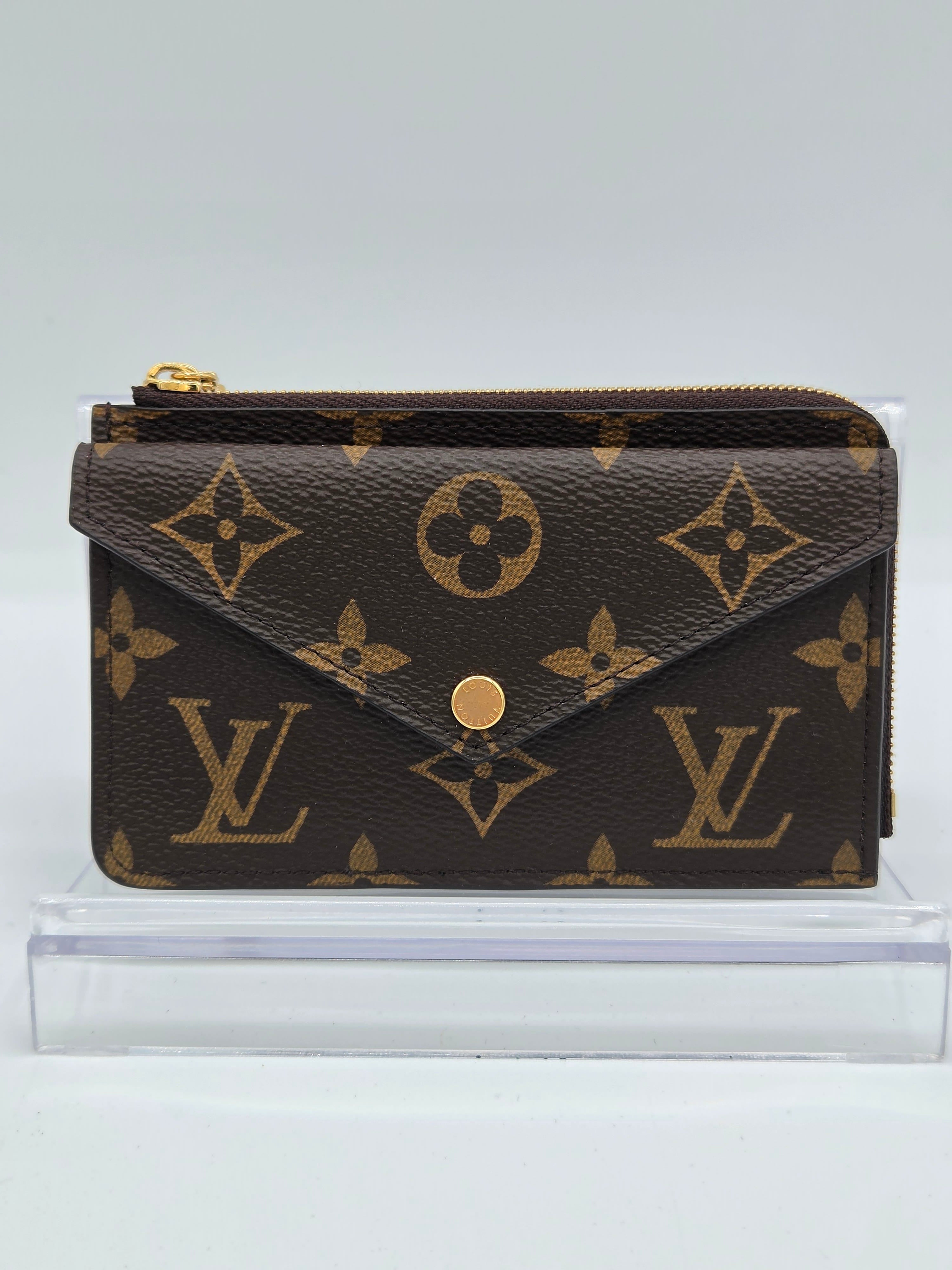 A Louis Vuitton monogram wallet in dark brown coated canvas with gold-tone hardware, shown standing upright next to a clear acrylic block against a light gray background.
