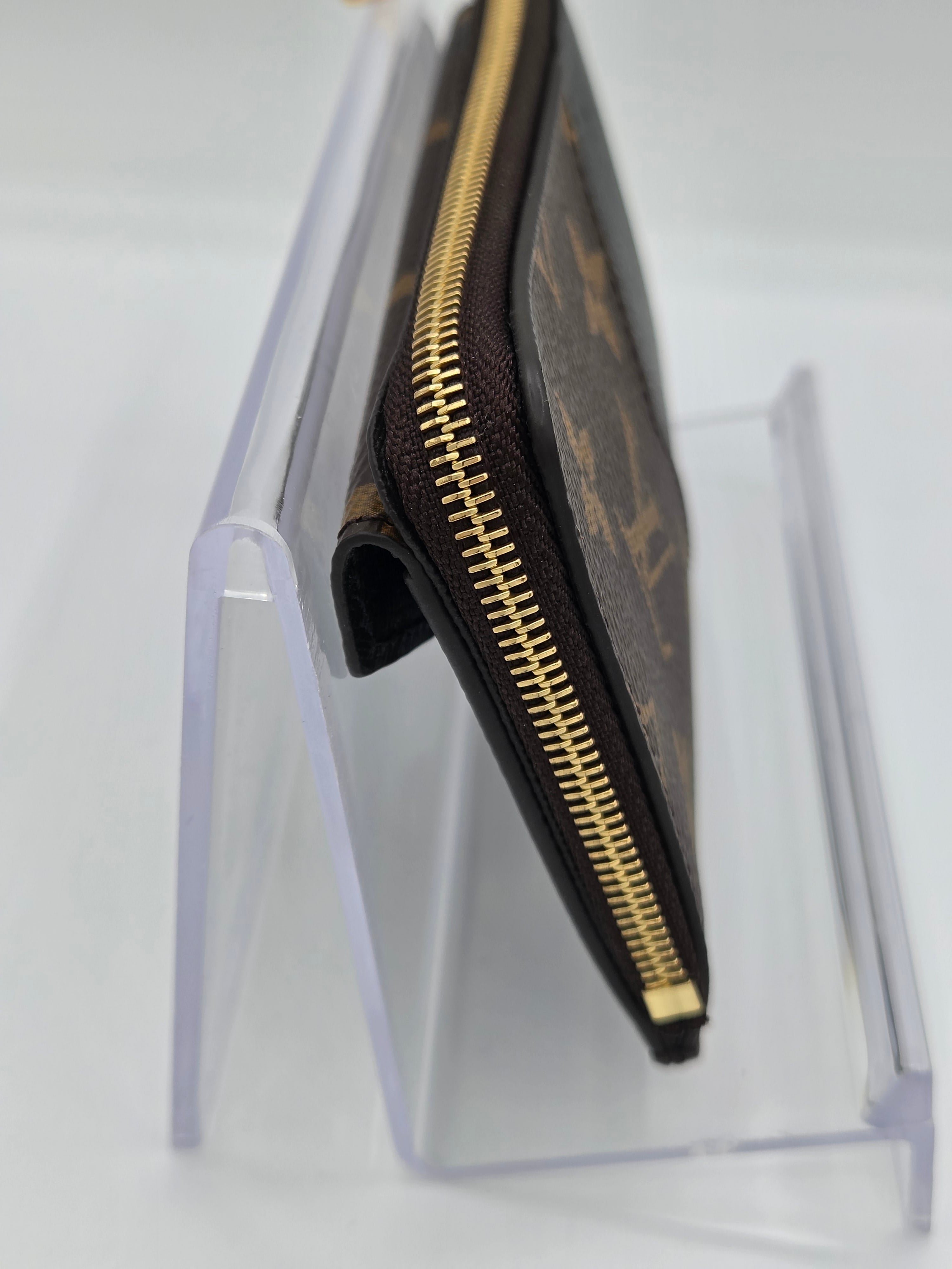 A brown monogram canvas zip-around wallet with gold-tone zippers, displayed on a clear acrylic stand against a white background.