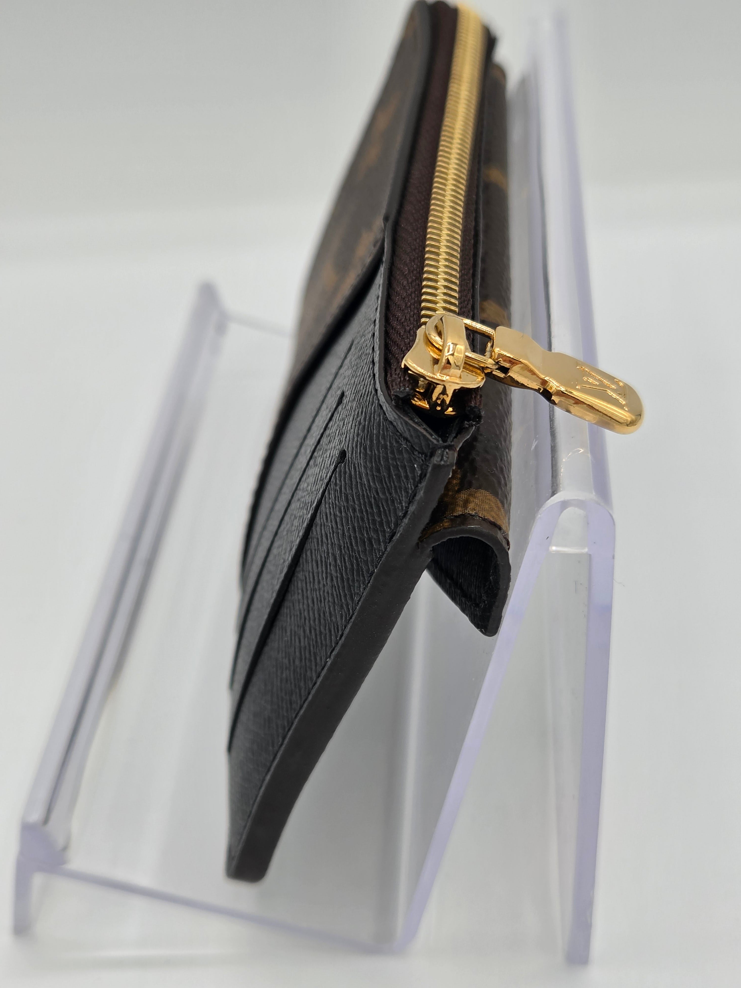 A Louis Vuitton monogram card holder wallet in dark brown and beige canvas, featuring a gold-tone zipper and a gold-tone logo tab, displayed on a clear acrylic stand.