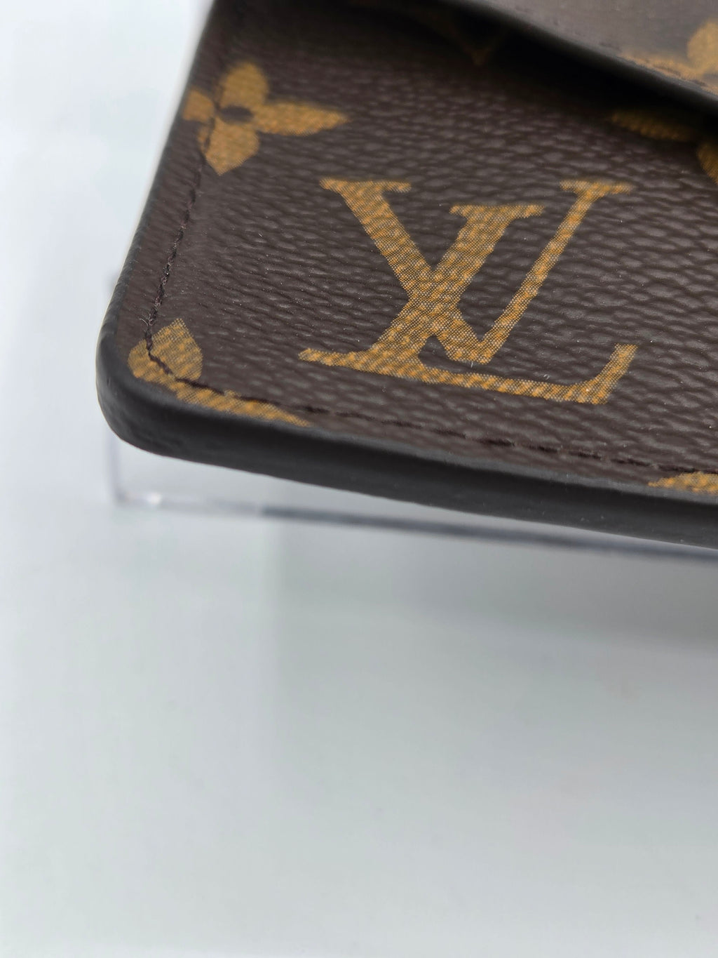 Close-up of a brown monogram canvas item with gold Louis Vuitton LV logo and floral motifs, showing stitched edges and a black trim against a light gray background.