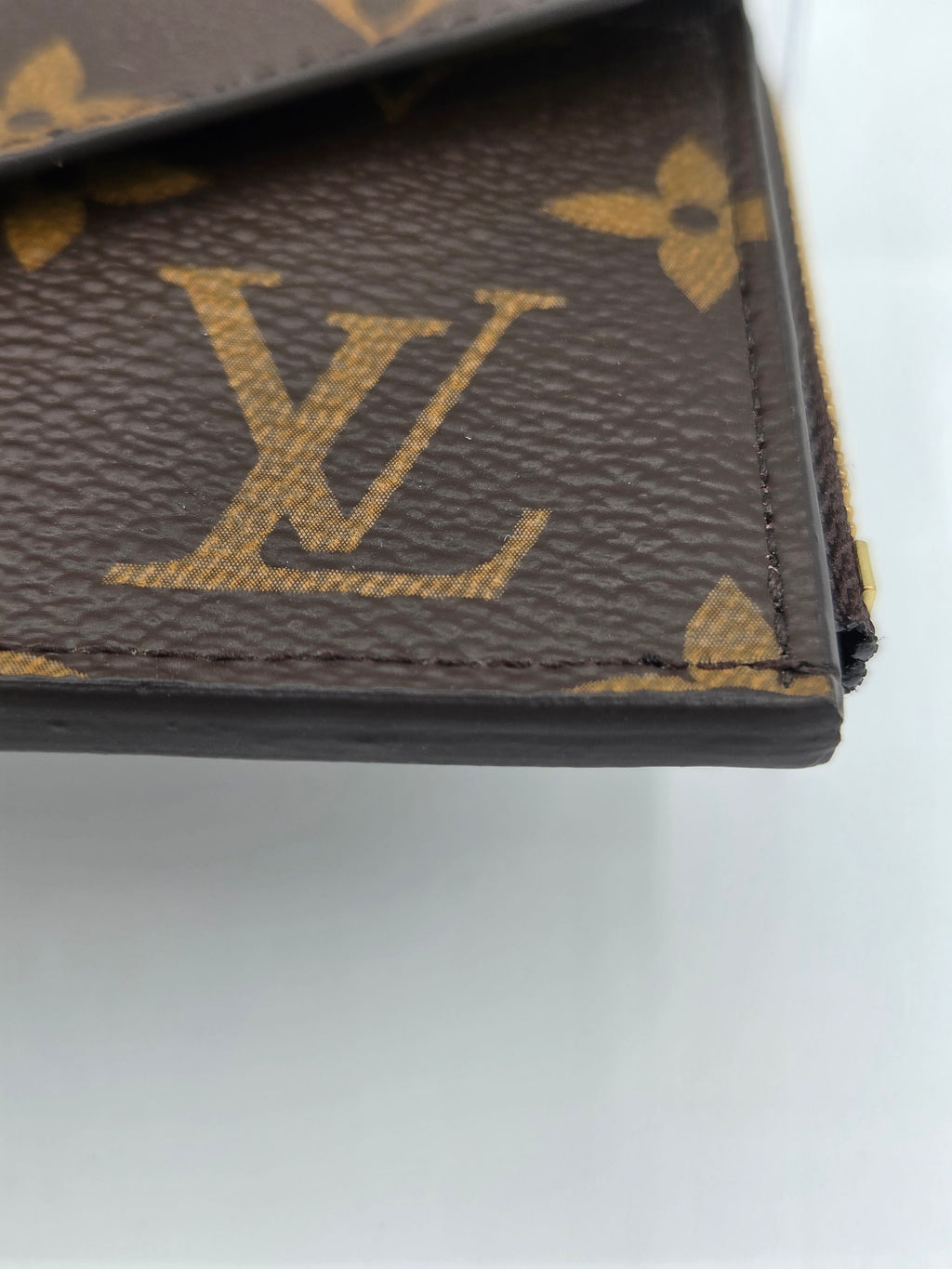 Close-up of a Louis Vuitton monogram wallet or card holder corner, showing the brown coated canvas with the signature tan/gold LV logo and floral print, along with dark brown leather edging.
