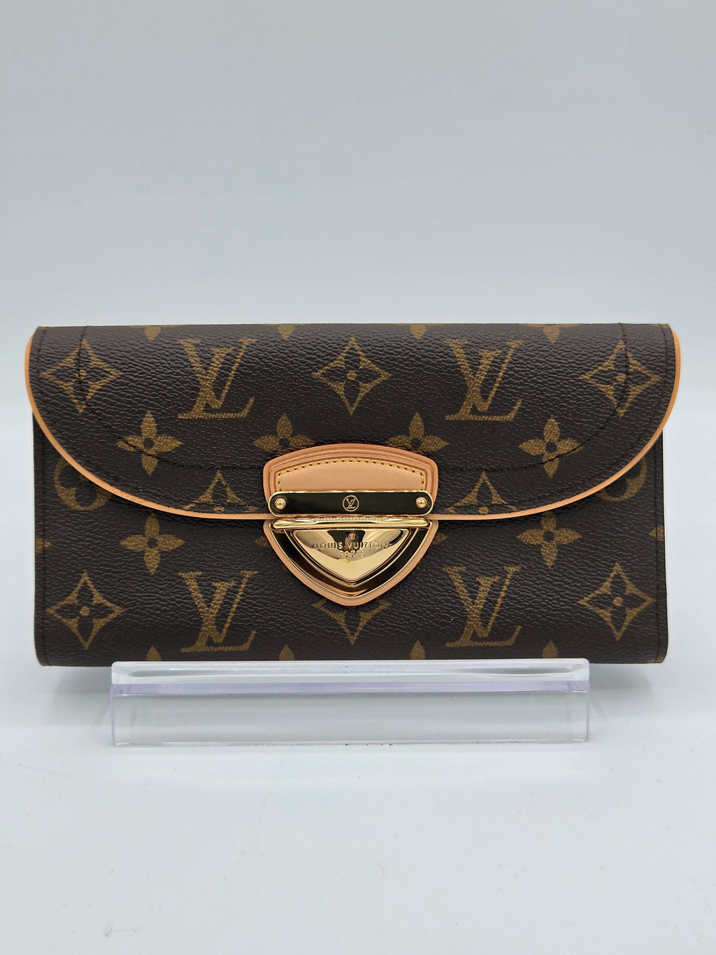 A Louis Vuitton monogram wallet in brown coated canvas with tan leather trim and a gold-tone turn-lock clasp, shown standing upright next to a clear plastic protective sleeve on a light gray background.