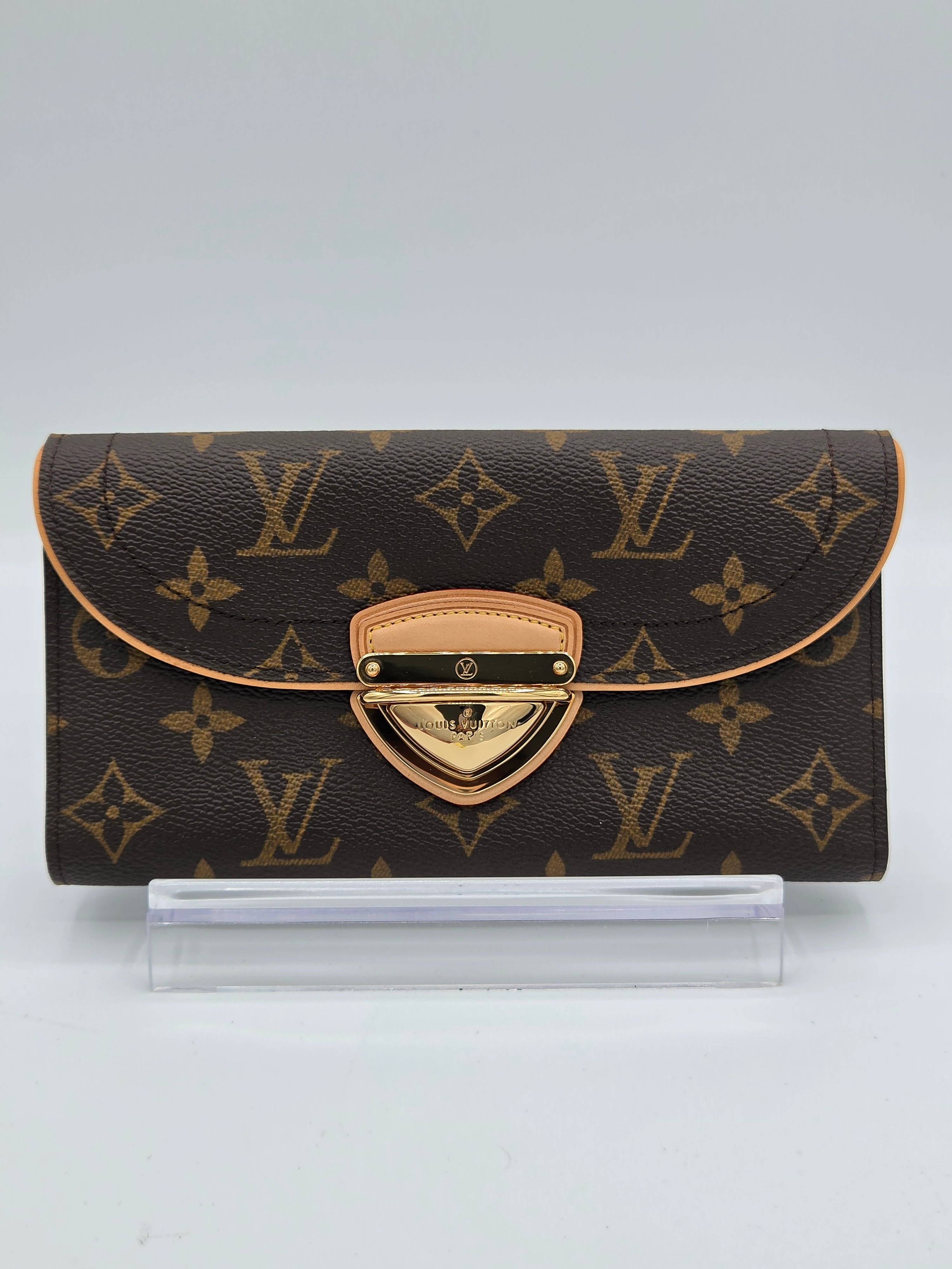 A Louis Vuitton monogram wallet in brown coated canvas with tan leather trim and a gold-tone turn-lock clasp, shown standing upright next to a clear plastic protective sleeve on a light gray background.