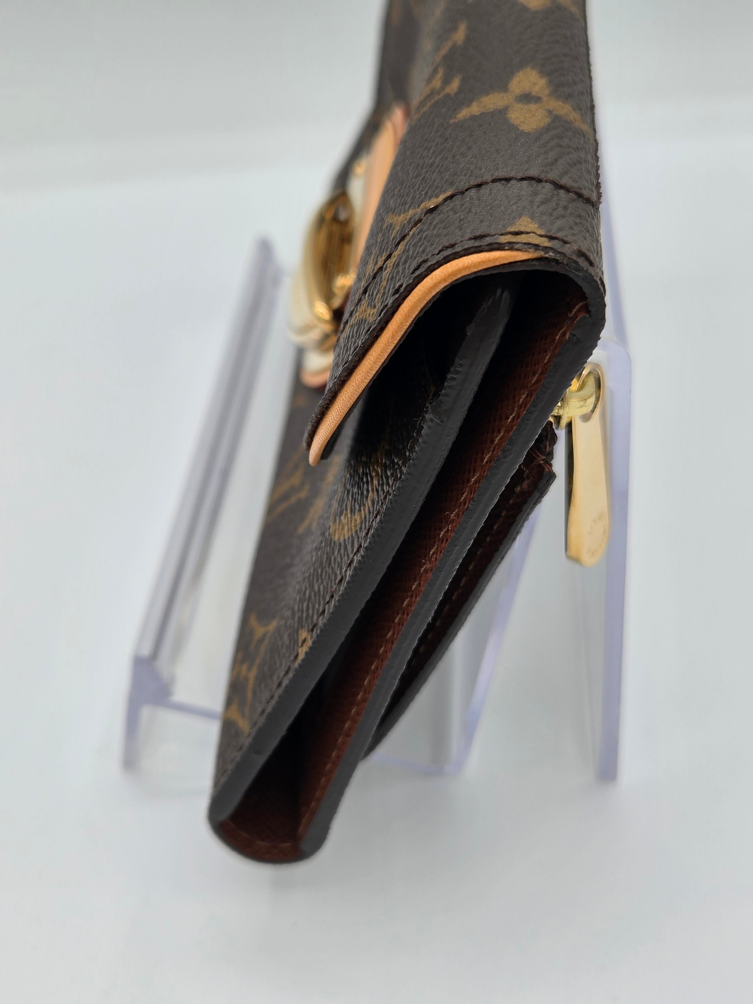 A close-up of a Louis Vuitton monogram wallet or card holder in brown coated canvas with gold-tone hardware, mounted on a clear display stand against a white background.