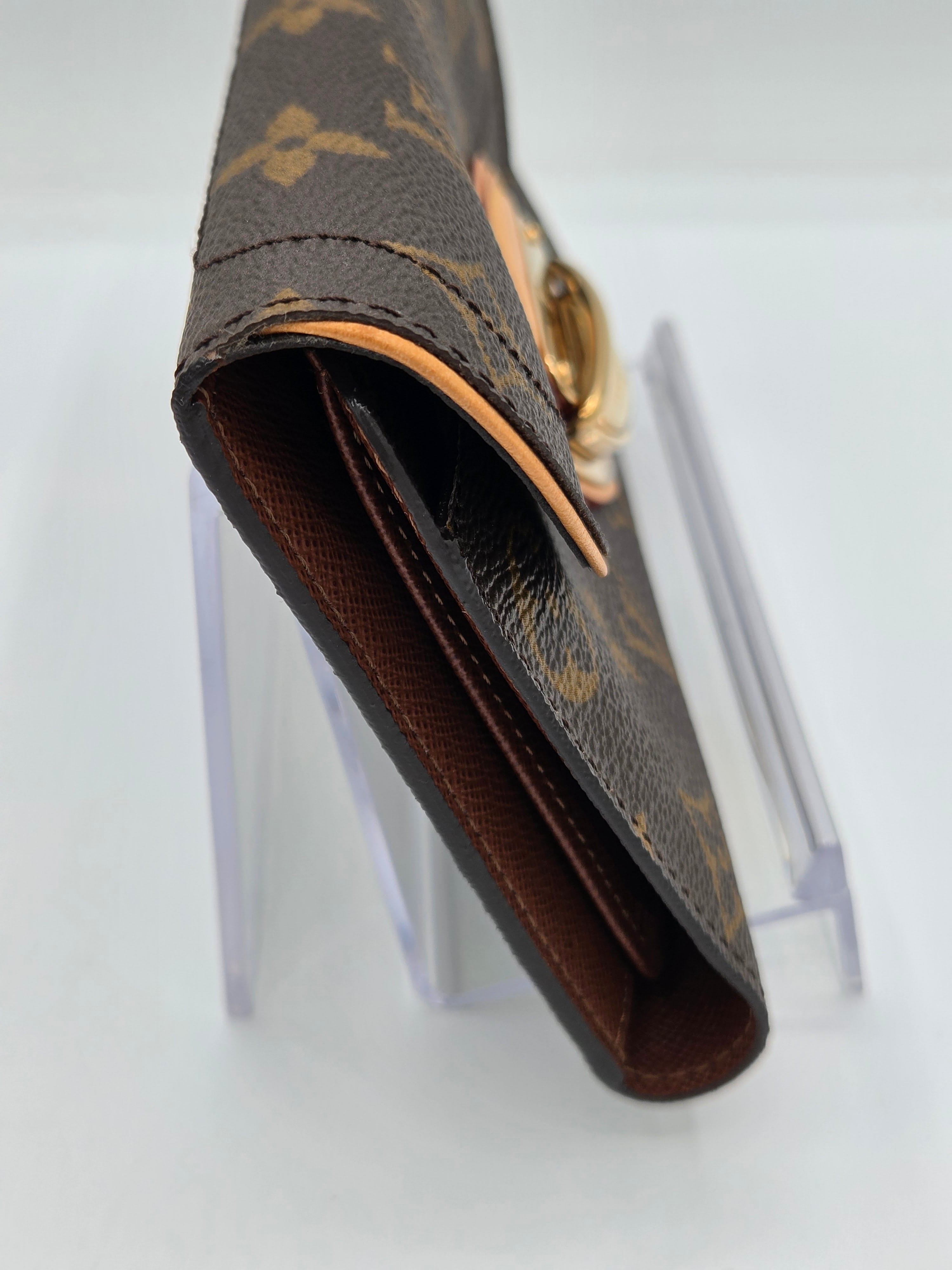 A close-up of a Louis Vuitton monogram wallet in brown coated canvas with gold-tone hardware, resting on a clear acrylic display stand against a white background.