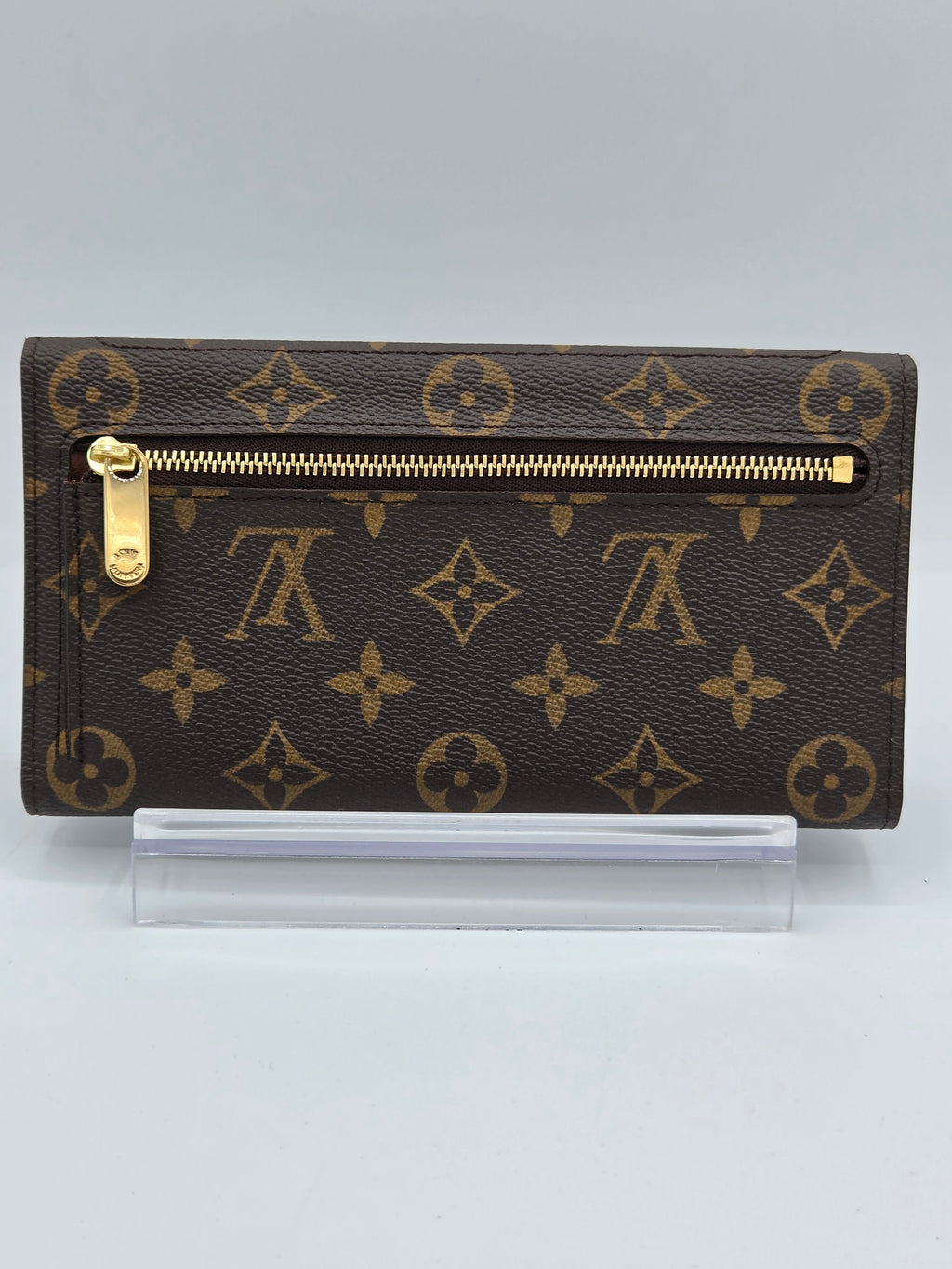 A Louis Vuitton monogram canvas zip-around wallet in dark brown with gold-tone hardware, shown upright next to a clear protective sleeve on a light gray background.