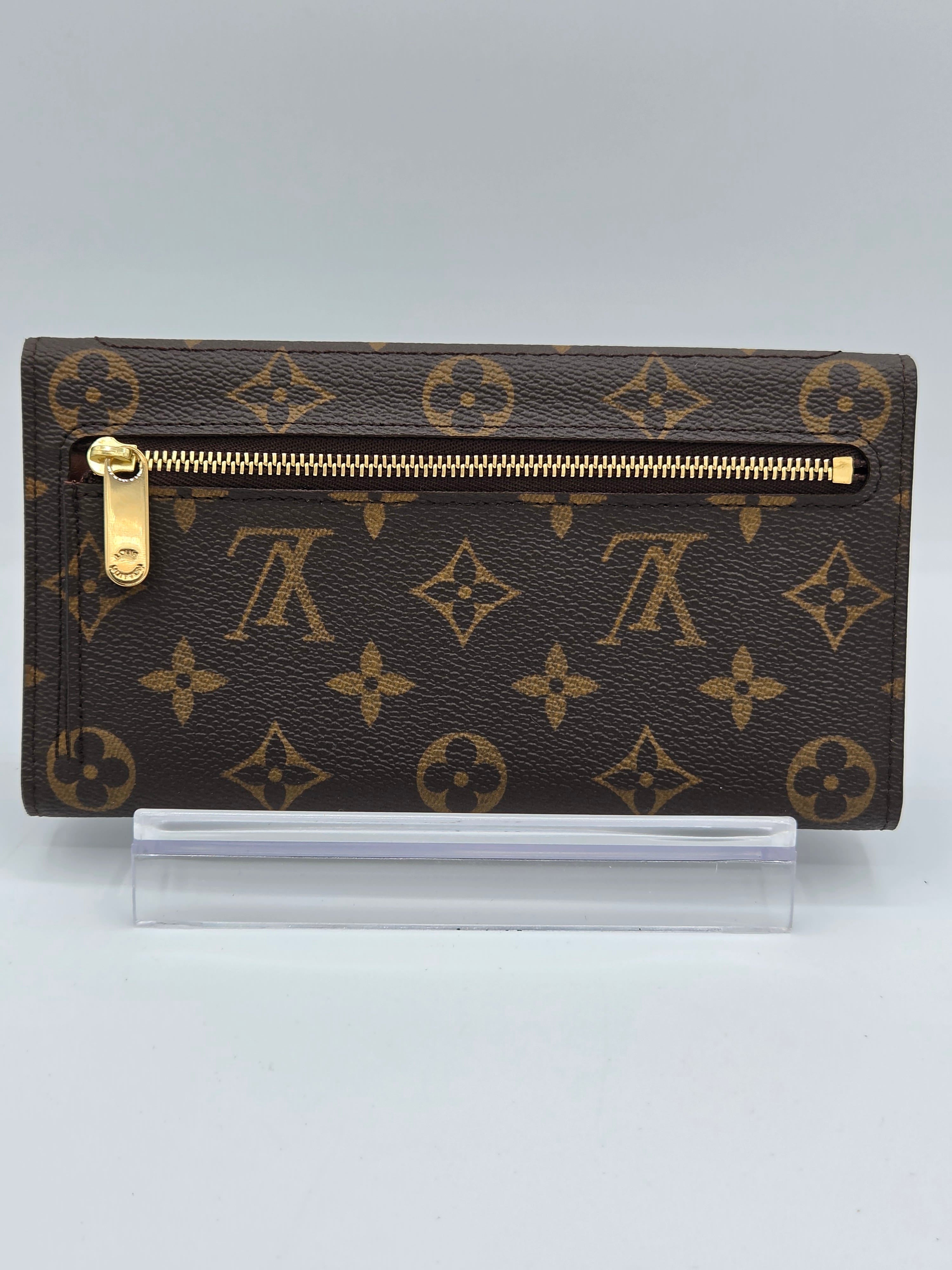A Louis Vuitton monogram canvas zip-around wallet in dark brown with gold-tone hardware, shown upright next to a clear protective sleeve on a light gray background.