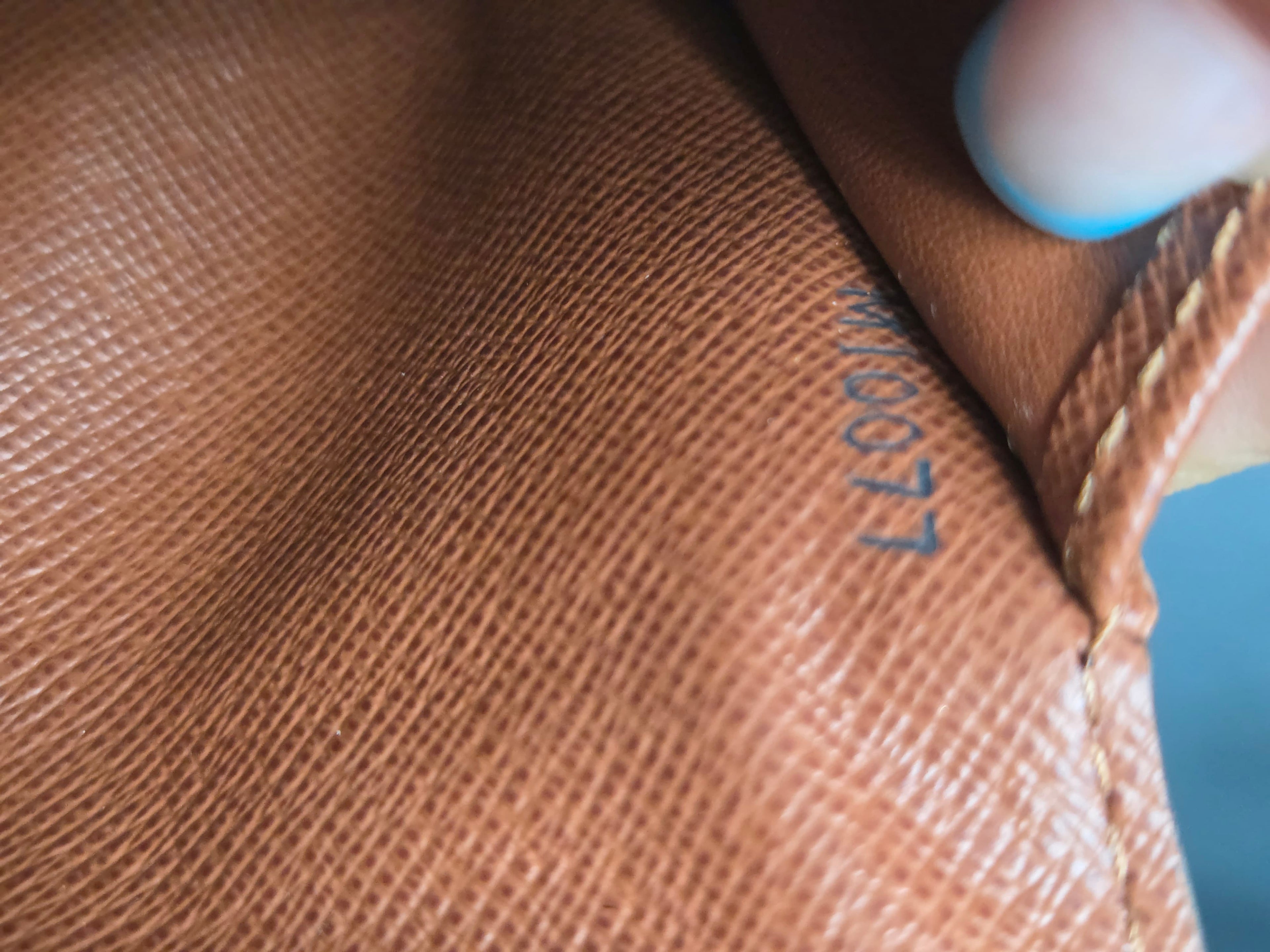 Close-up of a brown textured leather or canvas bag surface with a small dark printed alphanumeric mark and a finger holding the edge.
