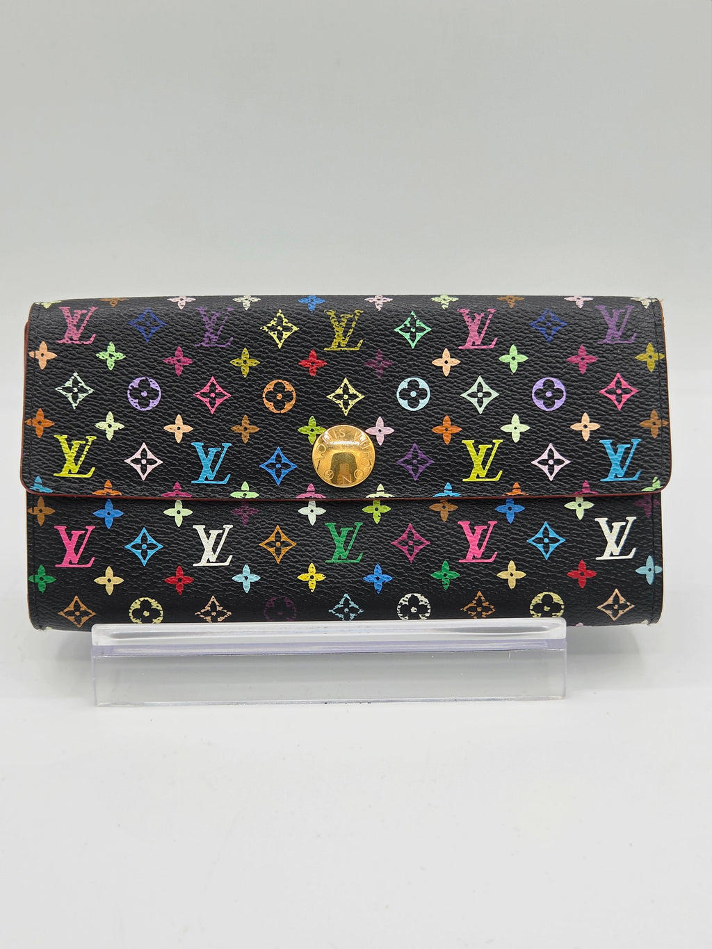 A Louis Vuitton long wallet in black monogram canvas with a vibrant multicolor LV pattern and a gold-tone turn-lock closure, displayed on a clear acrylic stand against a neutral gray background.