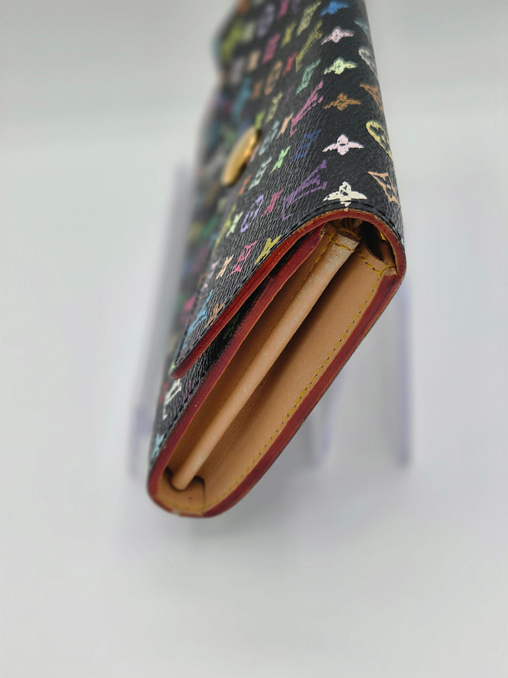 A close-up of a Louis Vuitton long wallet in black multicolor monogram canvas, showing the tan leather interior and red-brown edge trim, displayed on a white stand against a white background.
