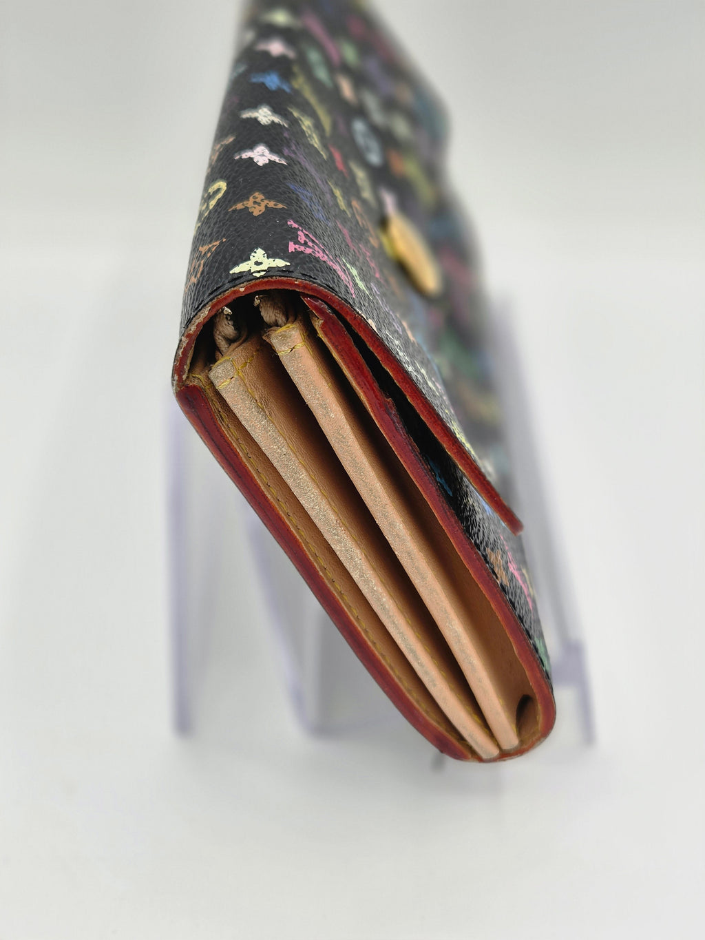 A close-up of a long wallet or clutch with a dark patterned exterior, red trim, and tan interior card slots, displayed on a clear stand against a white background.
