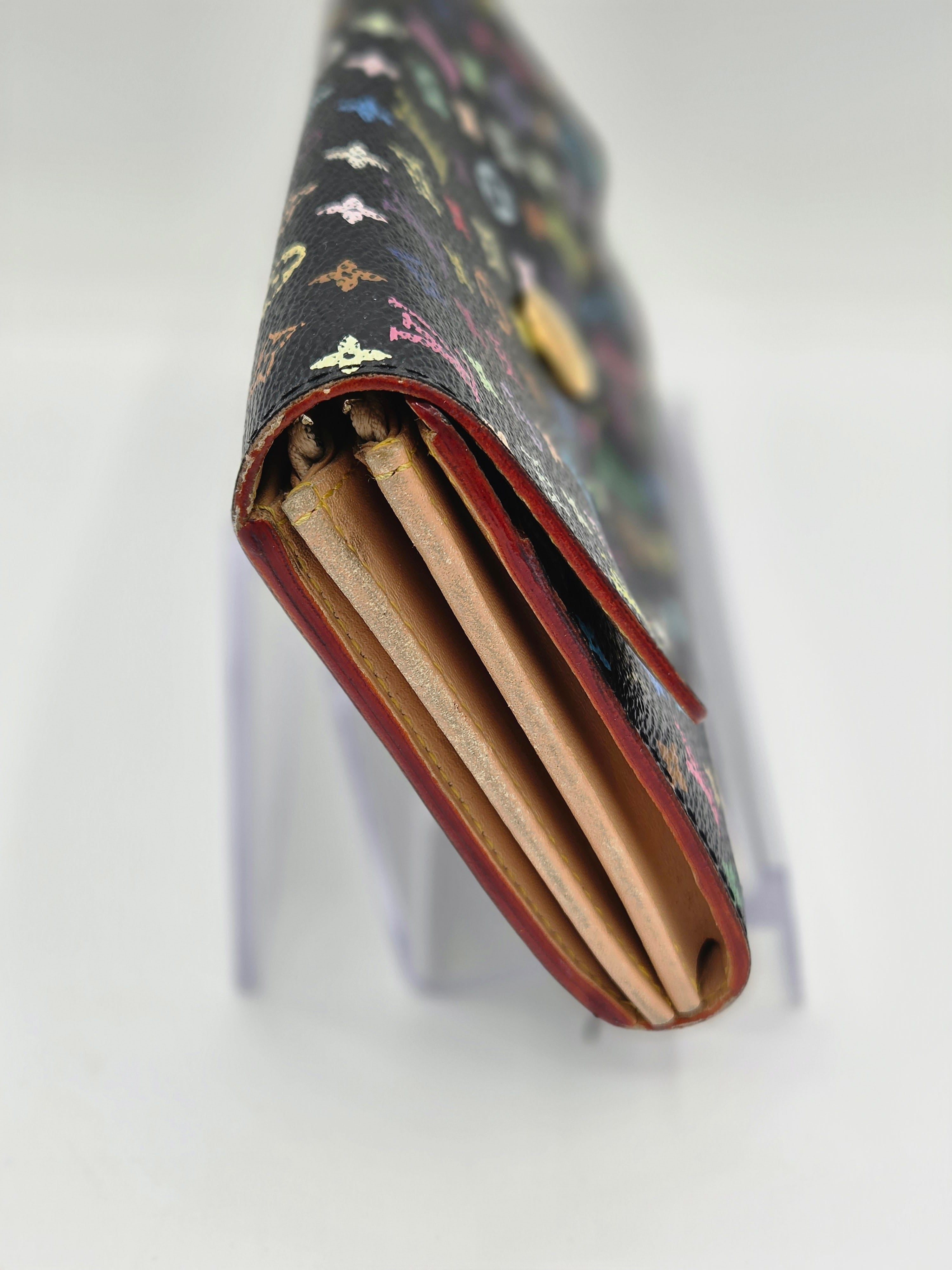 A close-up of a long wallet or clutch with a dark patterned exterior, red trim, and tan interior card slots, displayed on a clear stand against a white background.