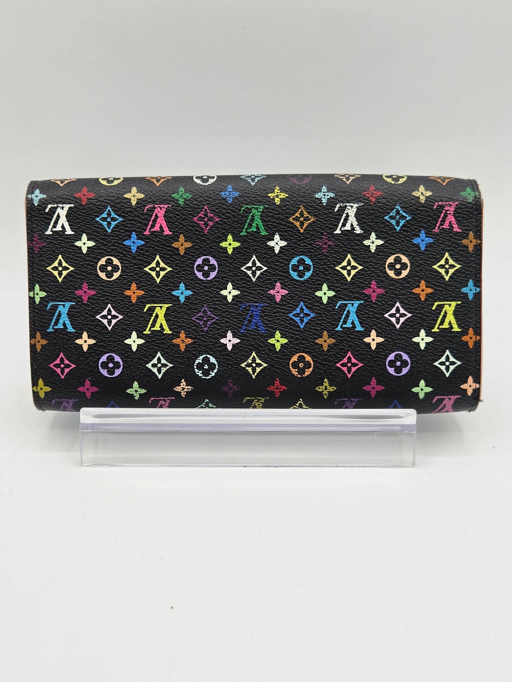 A Louis Vuitton monogram wallet in black coated canvas with a colorful repeating LV pattern, shown closed with a clear plastic sleeve on a light gray background.