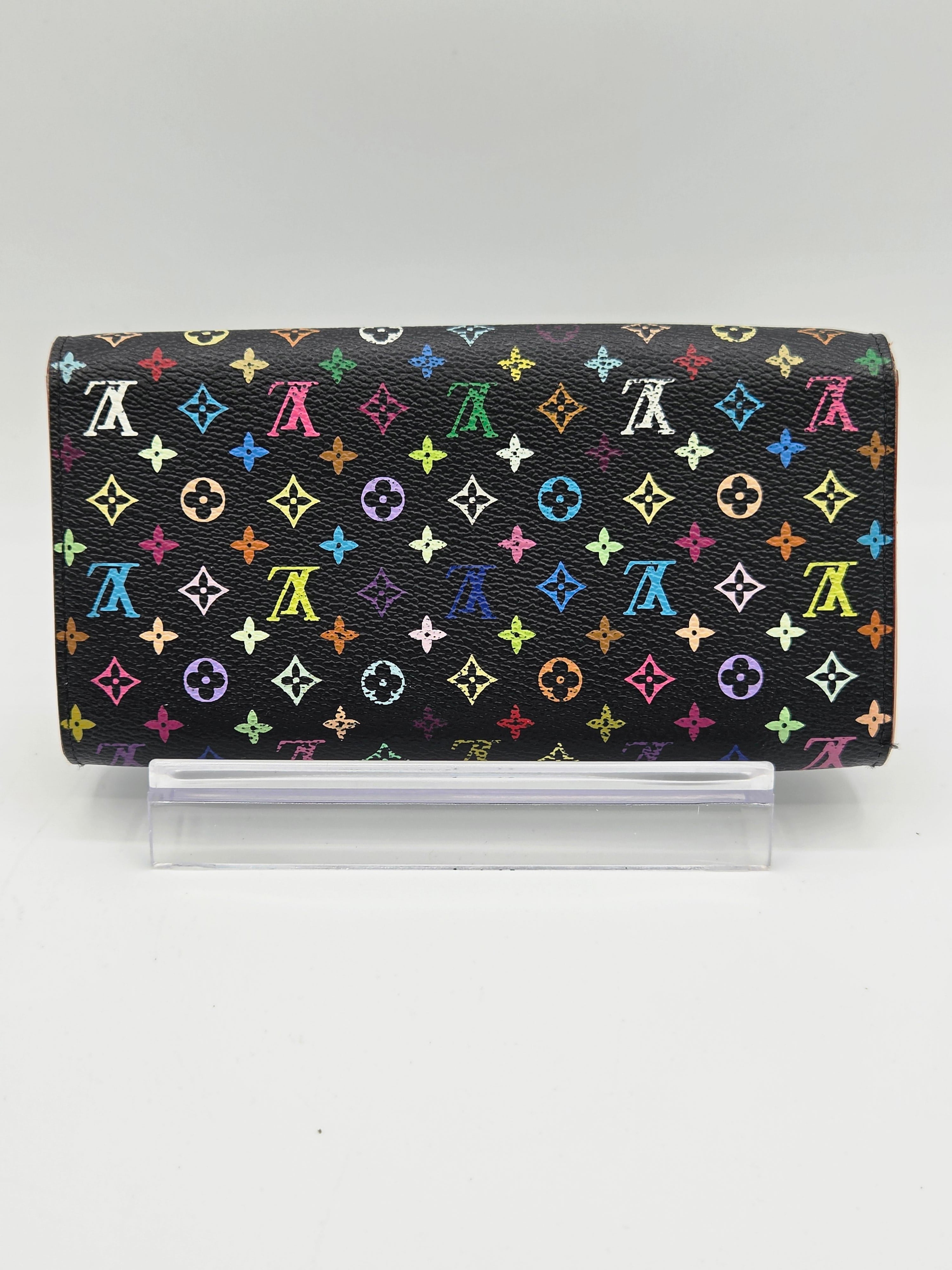 A Louis Vuitton monogram wallet in black coated canvas with a colorful repeating LV pattern, shown closed with a clear plastic sleeve on a light gray background.