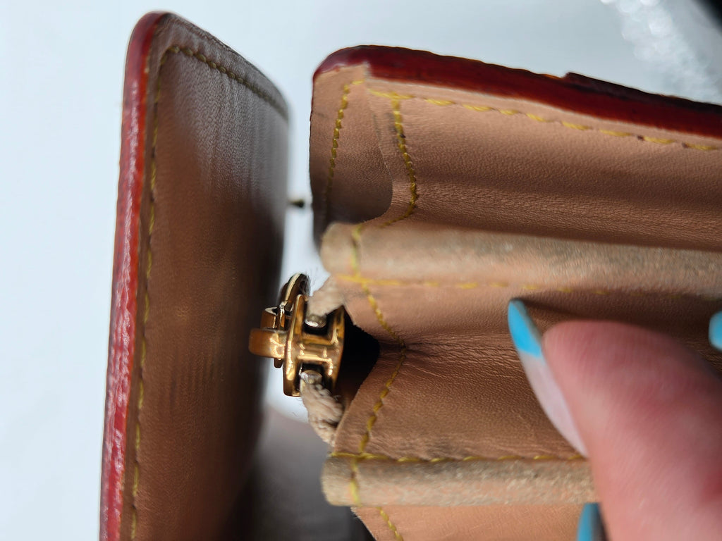 Close-up of an open brown leather wallet or card case showing tan interior lining, yellow stitching, a gold-tone clasp, and a finger with blue nail polish touching the edge.